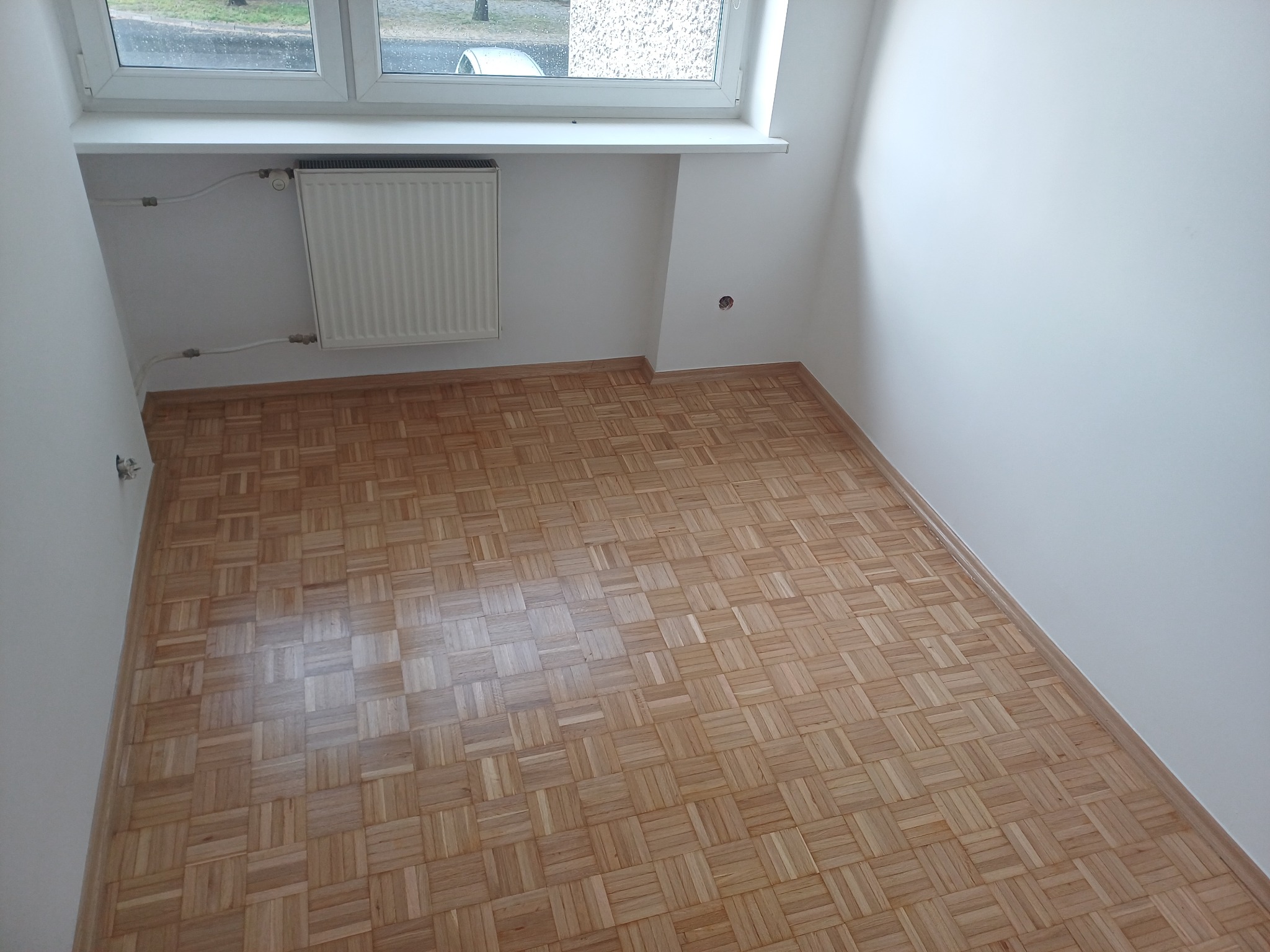 Parkiet w jodełkę po cyklinowaniu w pustym, jasnym pokoju z grzejnikiem pod oknem. Widoczne gładkie, drewniane klepki i świeżo pomalowane ściany.