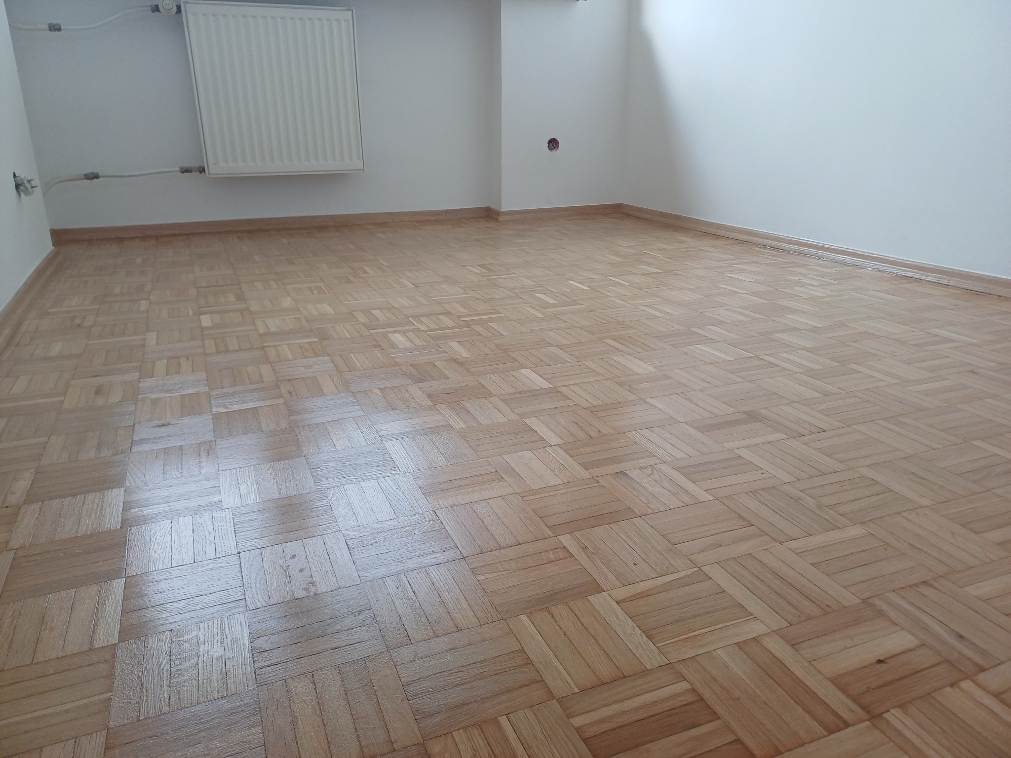 Parkiet po renowacji w pustym pokoju z grzejnikiem. Jasne drewno ułożone w kwadraty, widoczne listwy przypodłogowe i gładkie ściany. Świeżo wykończona podłoga.