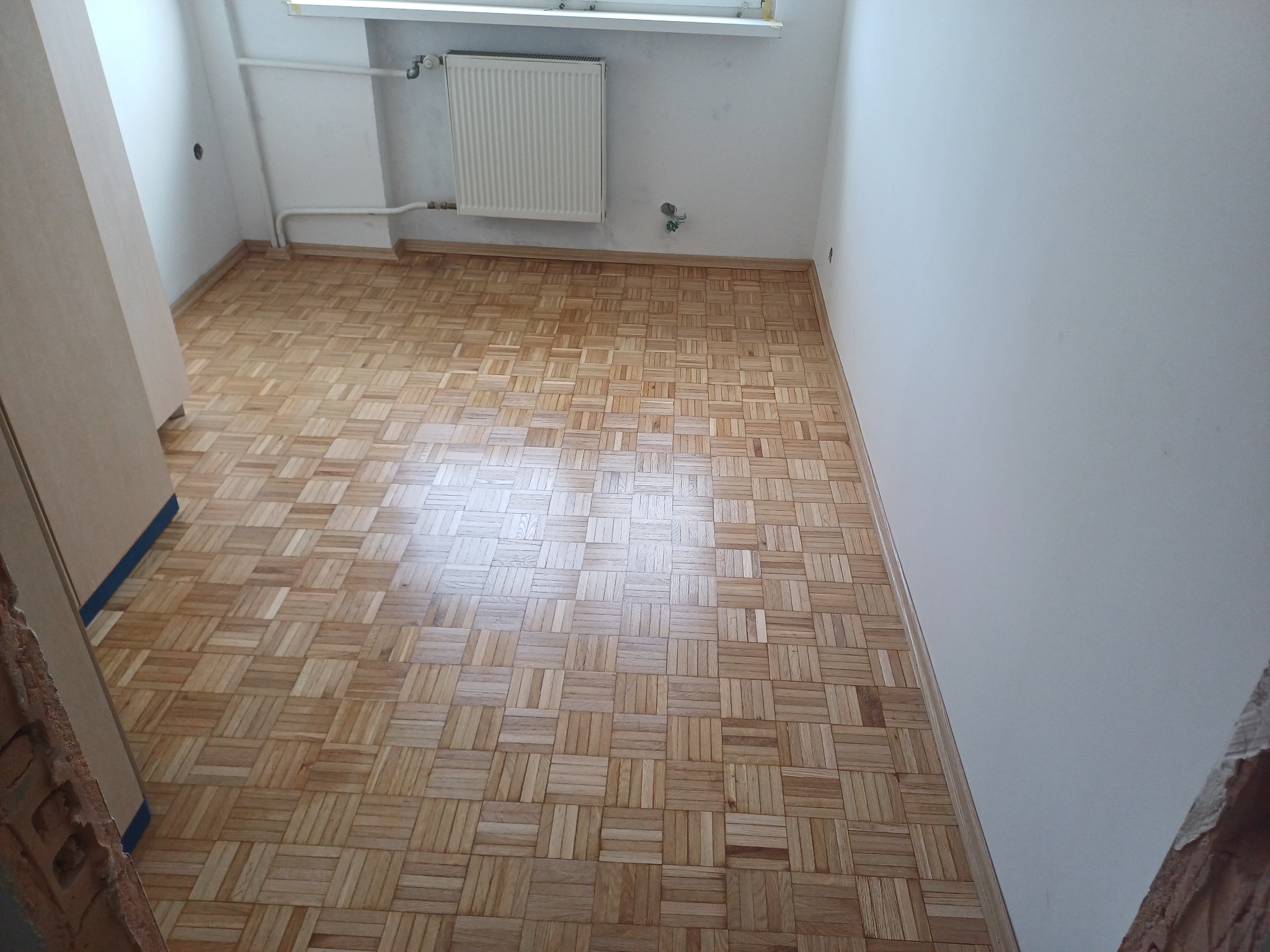 Parkiet ułożony w jodełkę w pustym pokoju z grzejnikiem. Widoczne ślady po remoncie na ścianach i brak listew przypodłogowych. Jasne drewno, naturalne światło.
