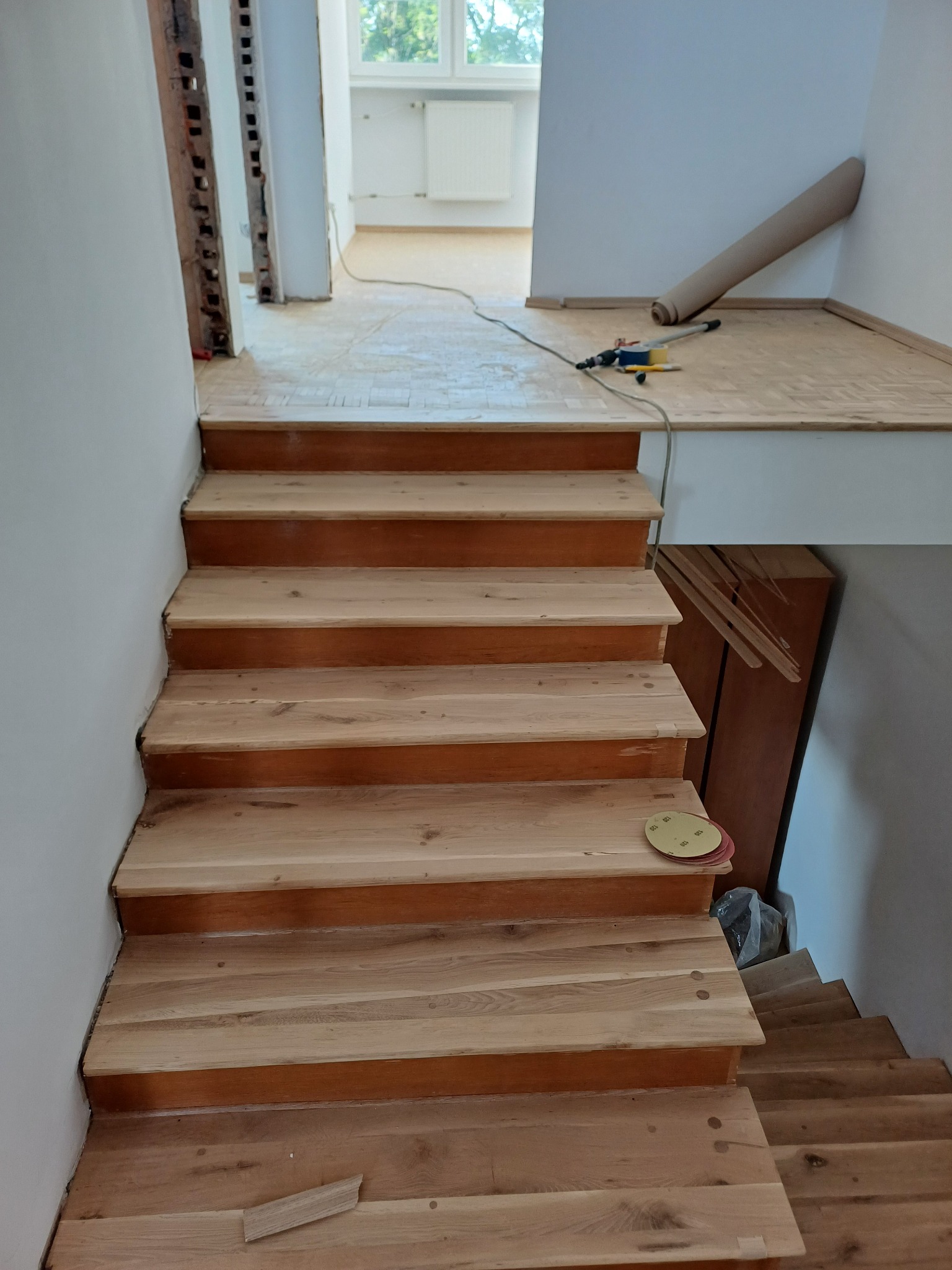Jasne, drewniane schody po renowacji, widoczne ślady prac wykończeniowych, w tle narzędzia i rolka papieru ściernego. Widok z góry na odnowione stopnie.