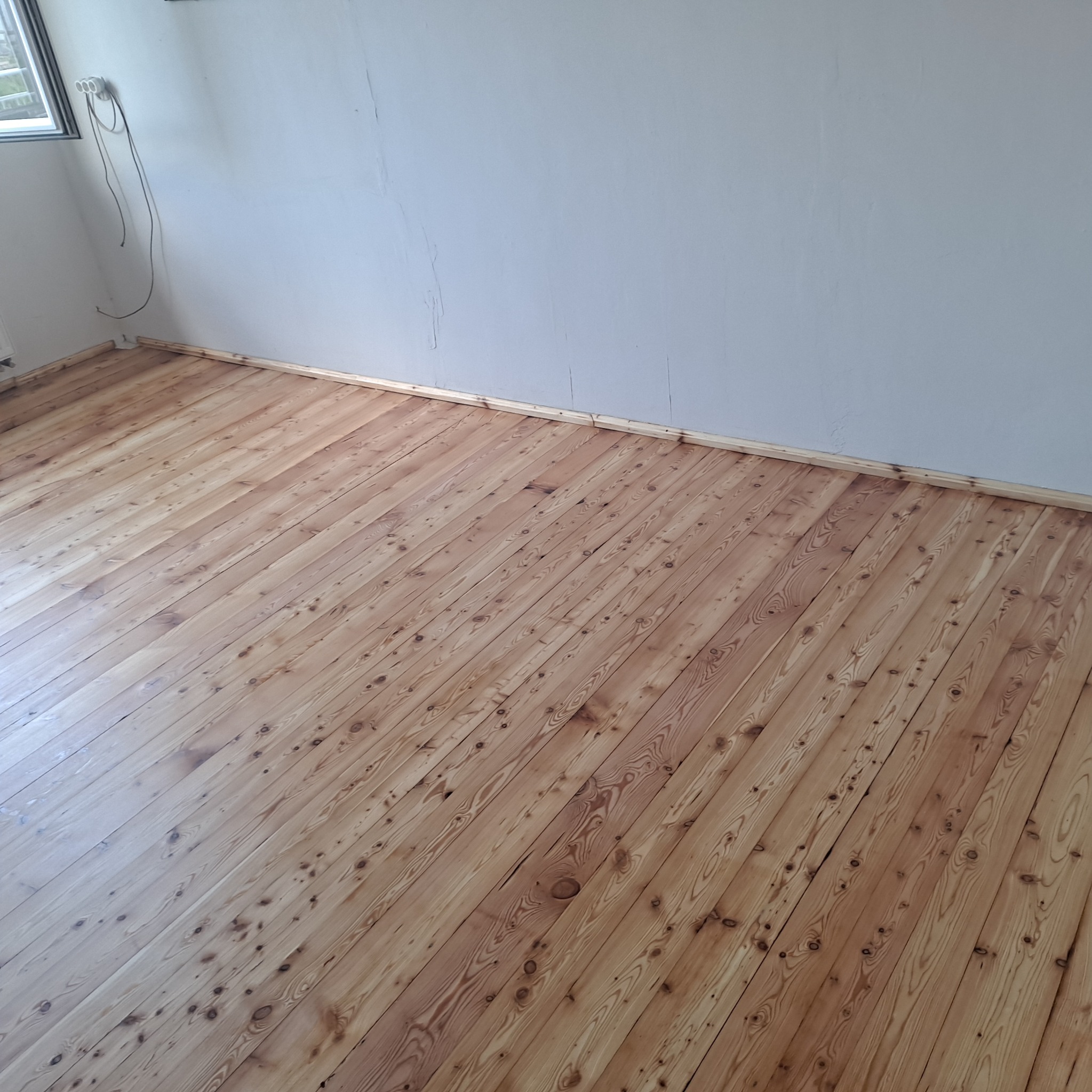 Jasna, odnowiona podłoga z desek w pokoju, widok z góry. Świeżo położone listwy przypodłogowe. Widoczne ślady po sękach i naturalny rysunek drewna.