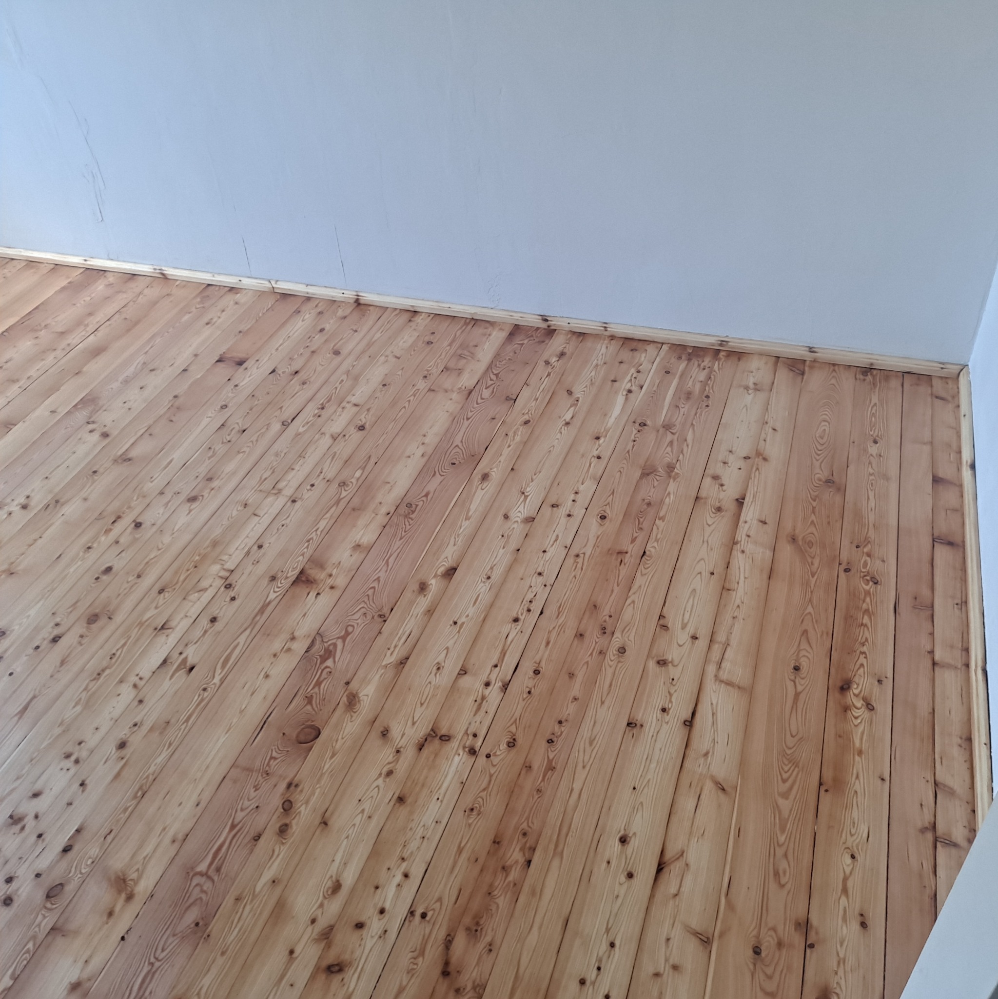 Jasna, odnowiona podłoga z desek ułożonych ukośnie, widoczna z perspektywy narożnika pokoju. Świeżo wycyklinowane drewno z wyraźnym usłojeniem i listwami przypodłogowymi.