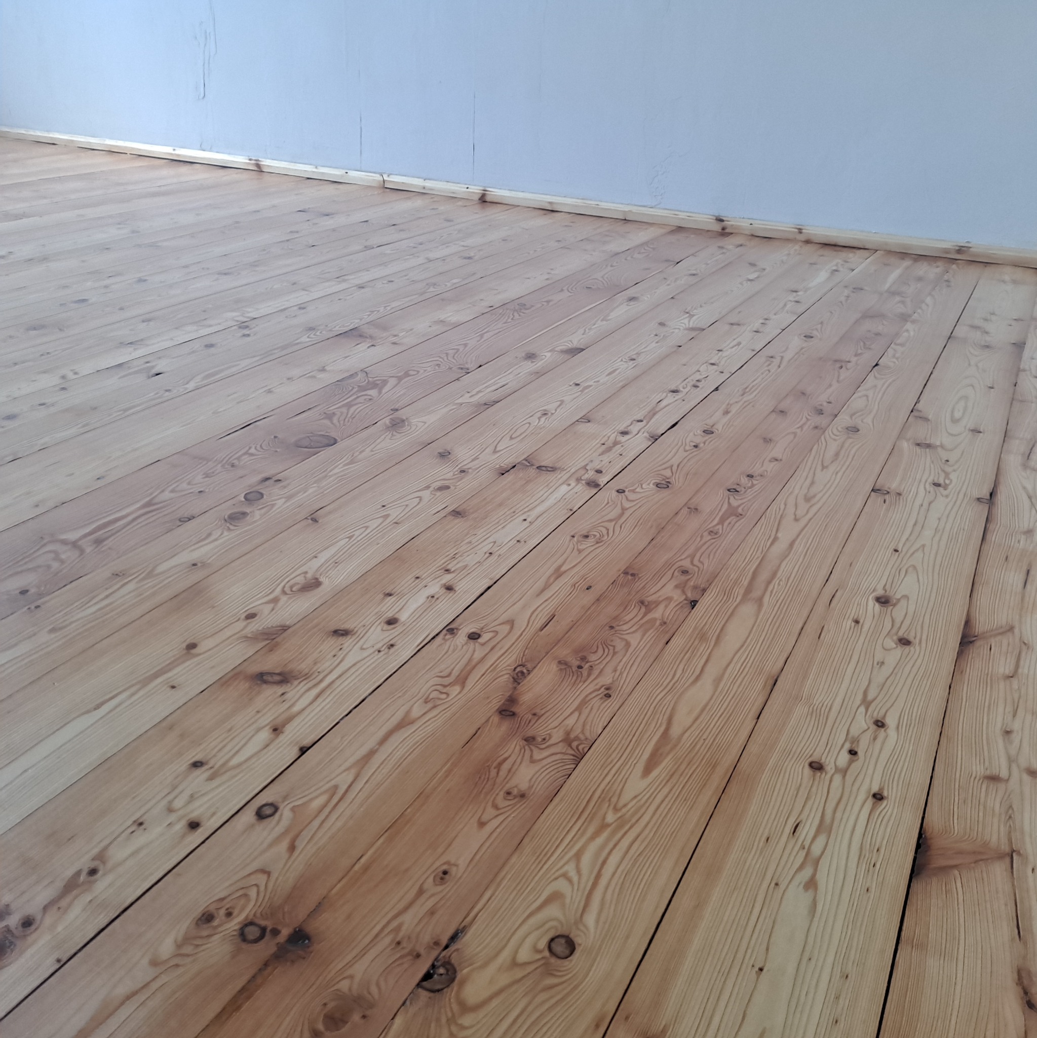 Jasna, odnowiona podłoga z desek sosnowych, widoczna z perspektywy pod kątem. Świeżo położona listwa przypodłogowa przy białej ścianie. Widoczne sęki i usłojenie drewna.