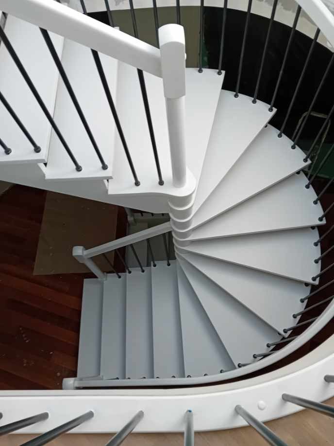 Białe, spiralne schody z czarnymi balustradami, widok z góry. Drewniana podłoga w tle. Minimalistyczny design, świeżo po renowacji. Jasne oświetlenie.