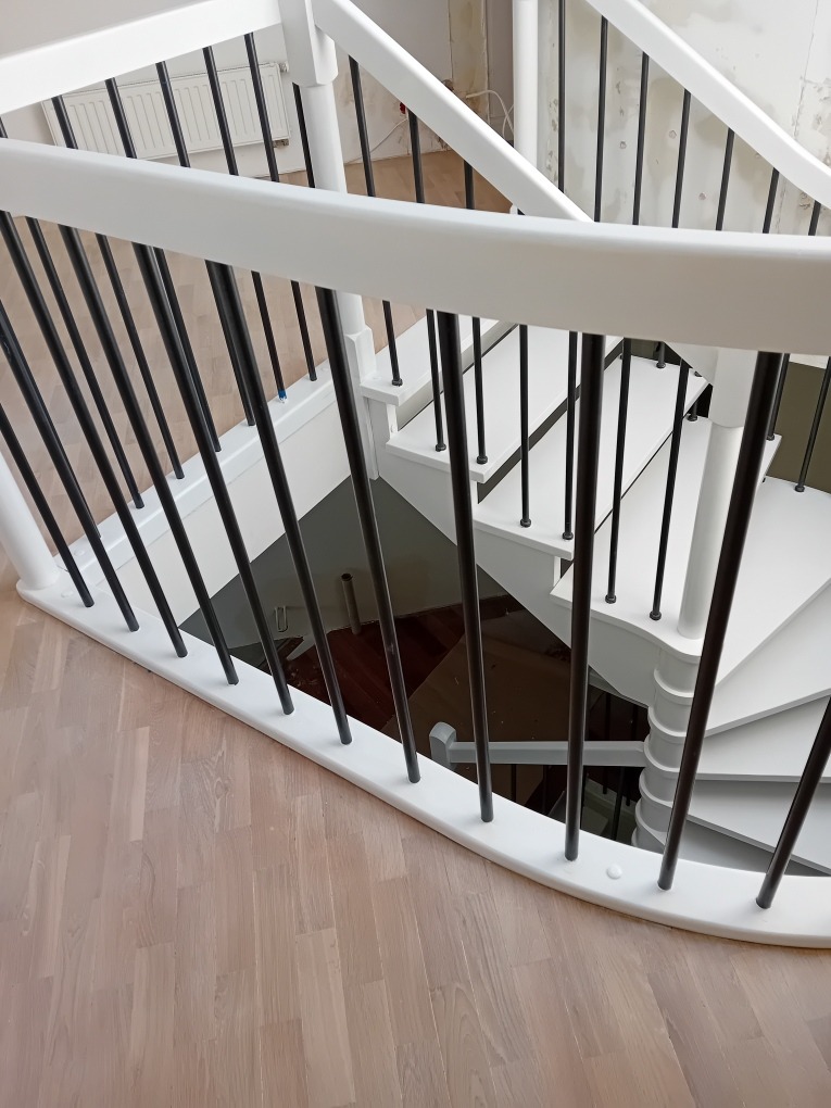 Białe, odnowione schody spiralne z czarnymi balustradami, widok z góry na drewnianą podłogę. Świeżo pomalowane, z widocznymi detalami wykończenia.