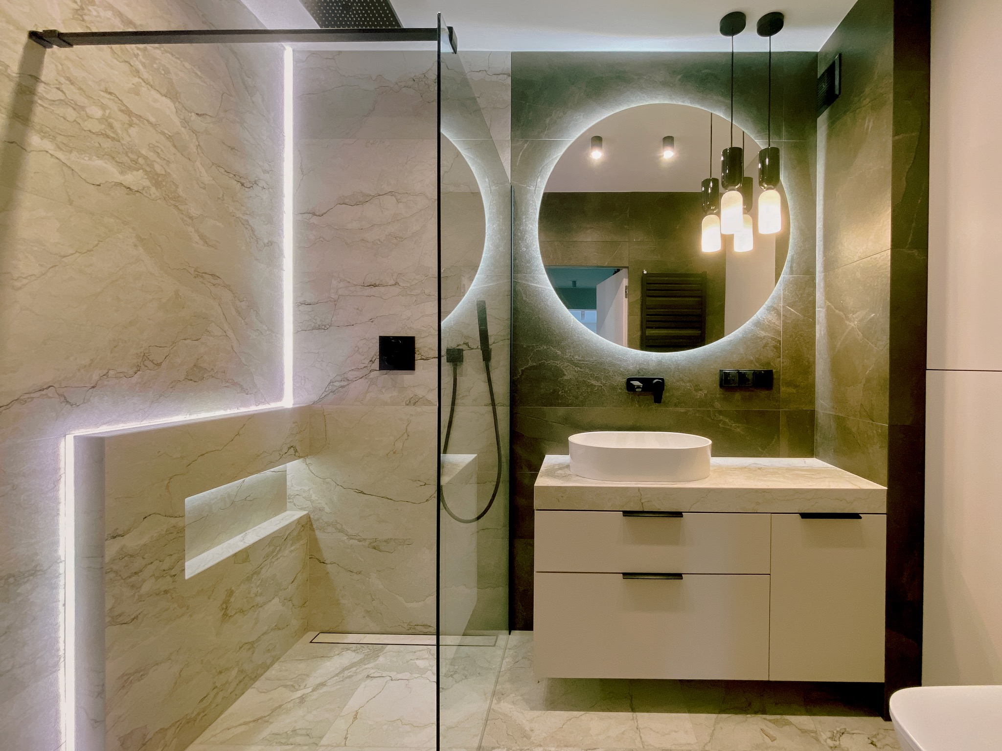 Elegancka łazienka z oświetleniem LED: kabina prysznicowa typu walk-in, okrągłe lustro z podświetleniem, umywalka nablatowa na szafce. Płytki imitujące marmur.