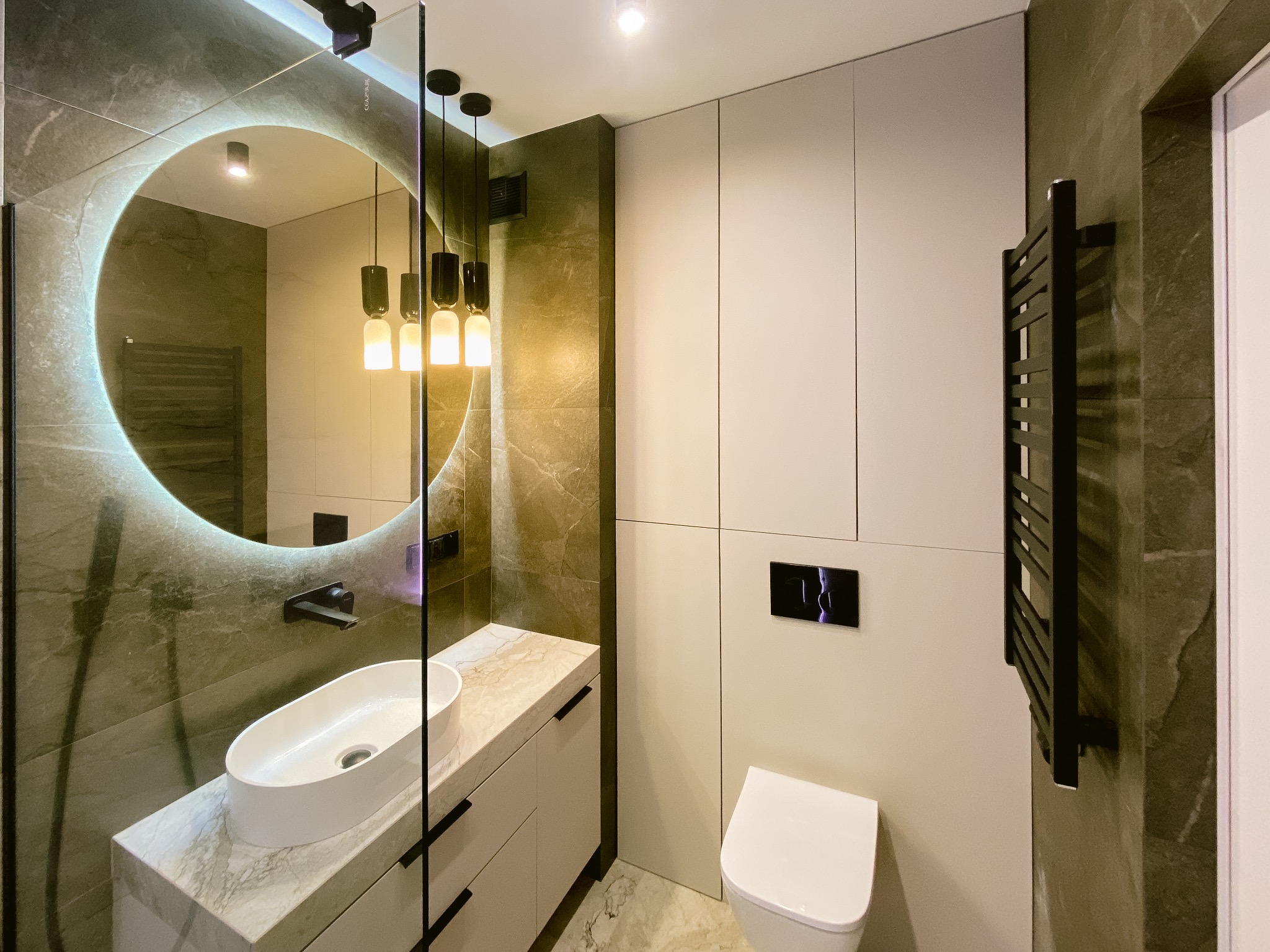 Nowoczesna łazienka z okrągłym lustrem z podświetleniem LED, umywalką nablatową na blacie z imitacji marmuru i czarnym grzejnikiem drabinkowym.