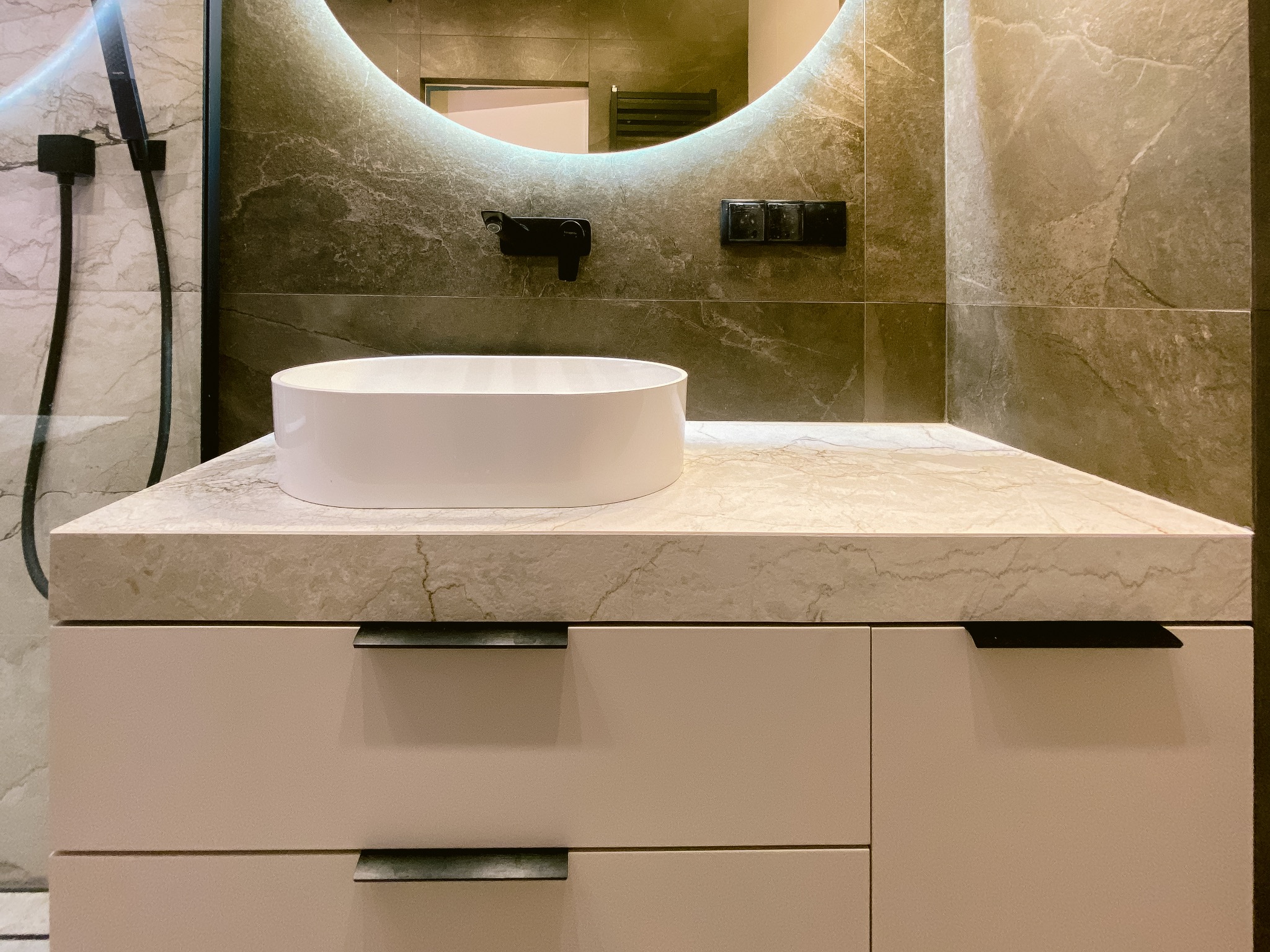 Nowoczesna łazienka z okrągłym lustrem LED, białą umywalką nablatową i szarymi płytkami imitującymi marmur. Minimalistyczne czarne akcenty armatury i włączników.