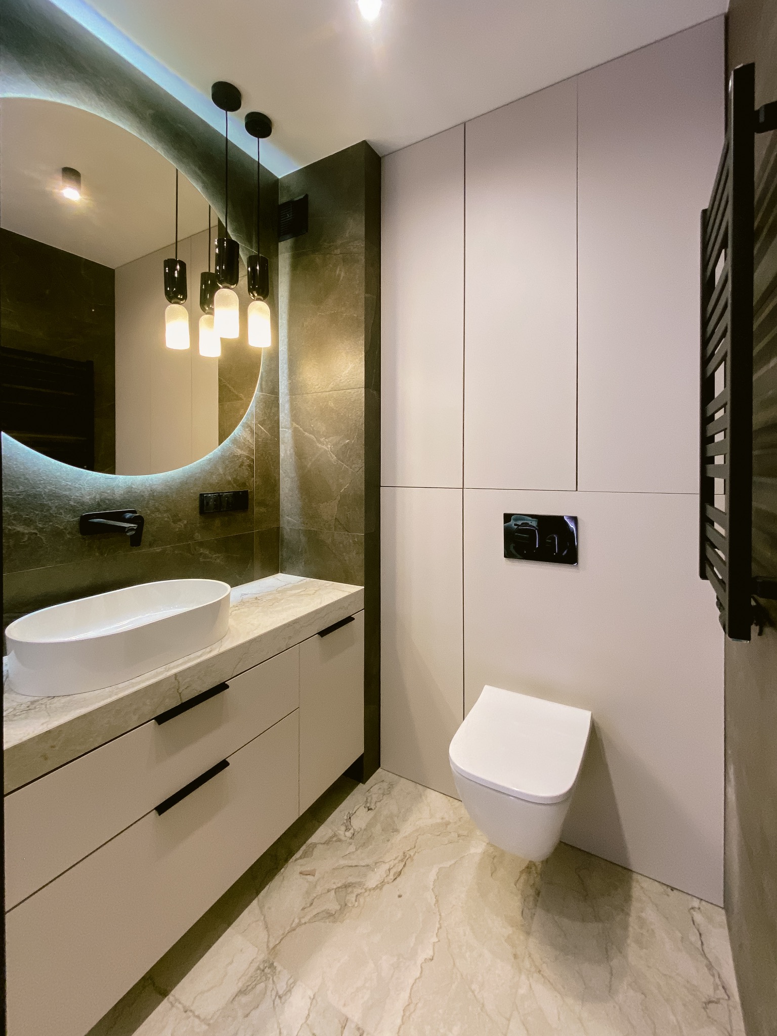 Nowoczesna łazienka z owalną umywalką, okrągłym lustrem z podświetleniem LED i wiszącą toaletą. Marmurowa podłoga i płytki w odcieniach szarości. Czarna armatura.