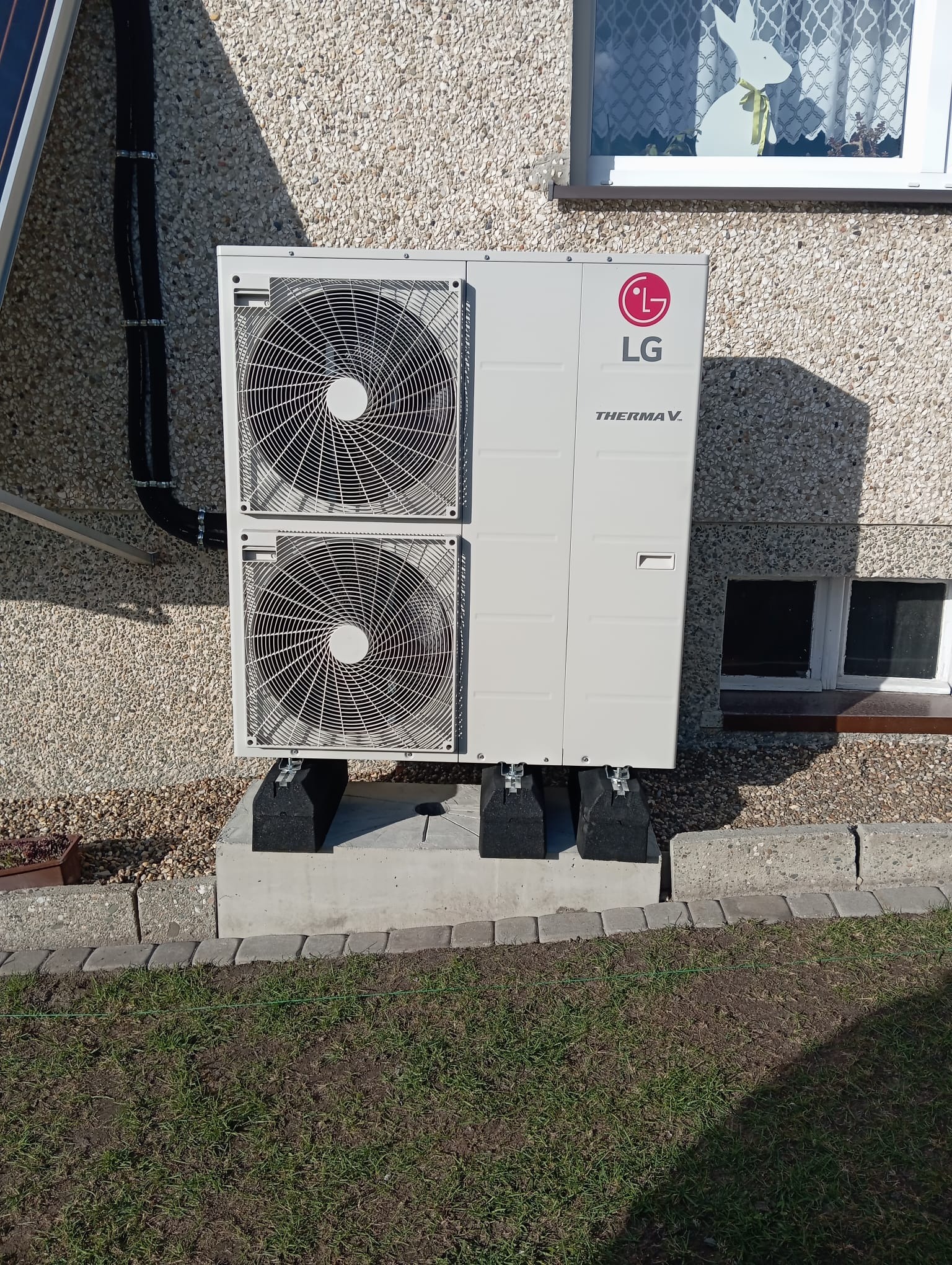 Biała pompa ciepła LG Therma V zamontowana na betonowym fundamencie przy ścianie budynku z oknem, w tle widoczny panel słoneczny.