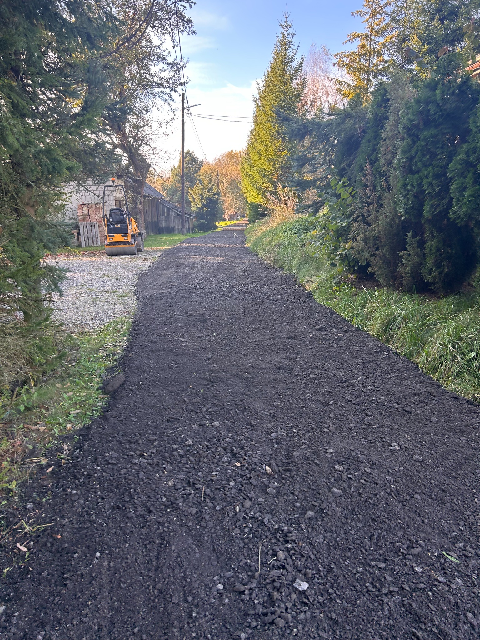 Utwardzona droga gruntowa z widocznym walcem drogowym w tle, otoczona zielenią i drzewami, w słoneczny dzień.