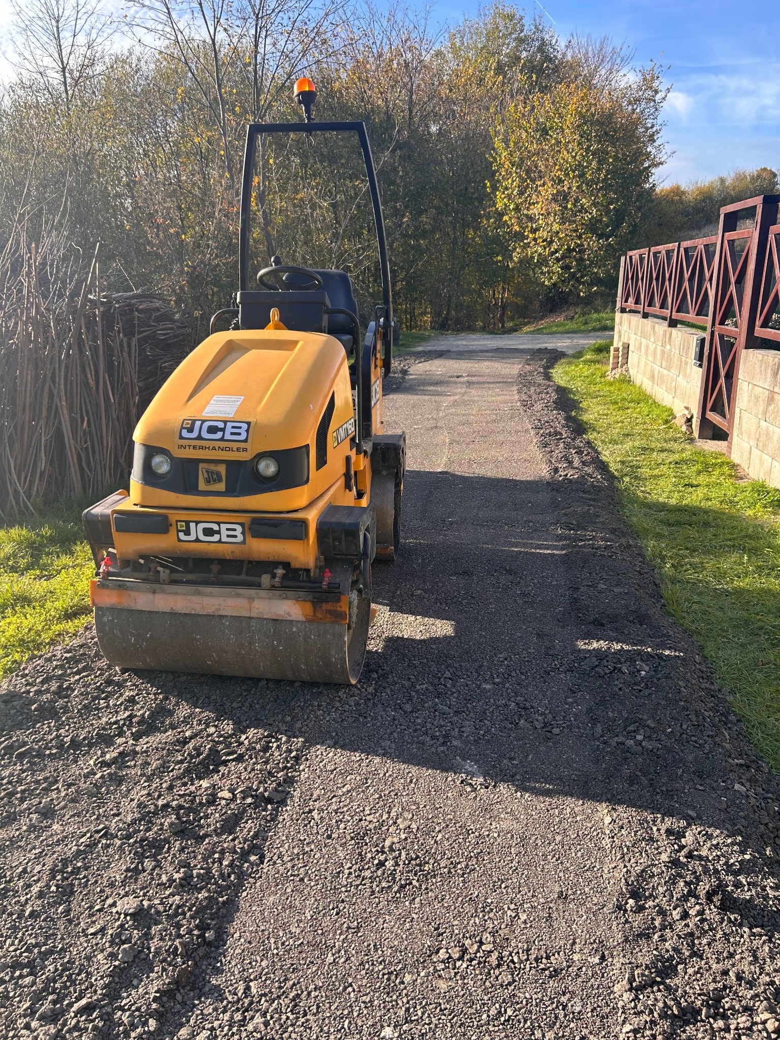 Żółty walec JCB ubija świeżo rozłożony asfalt na ścieżce, otoczony zielenią i drzewami. Widoczny fragment muru z metalowym ogrodzeniem w tle.