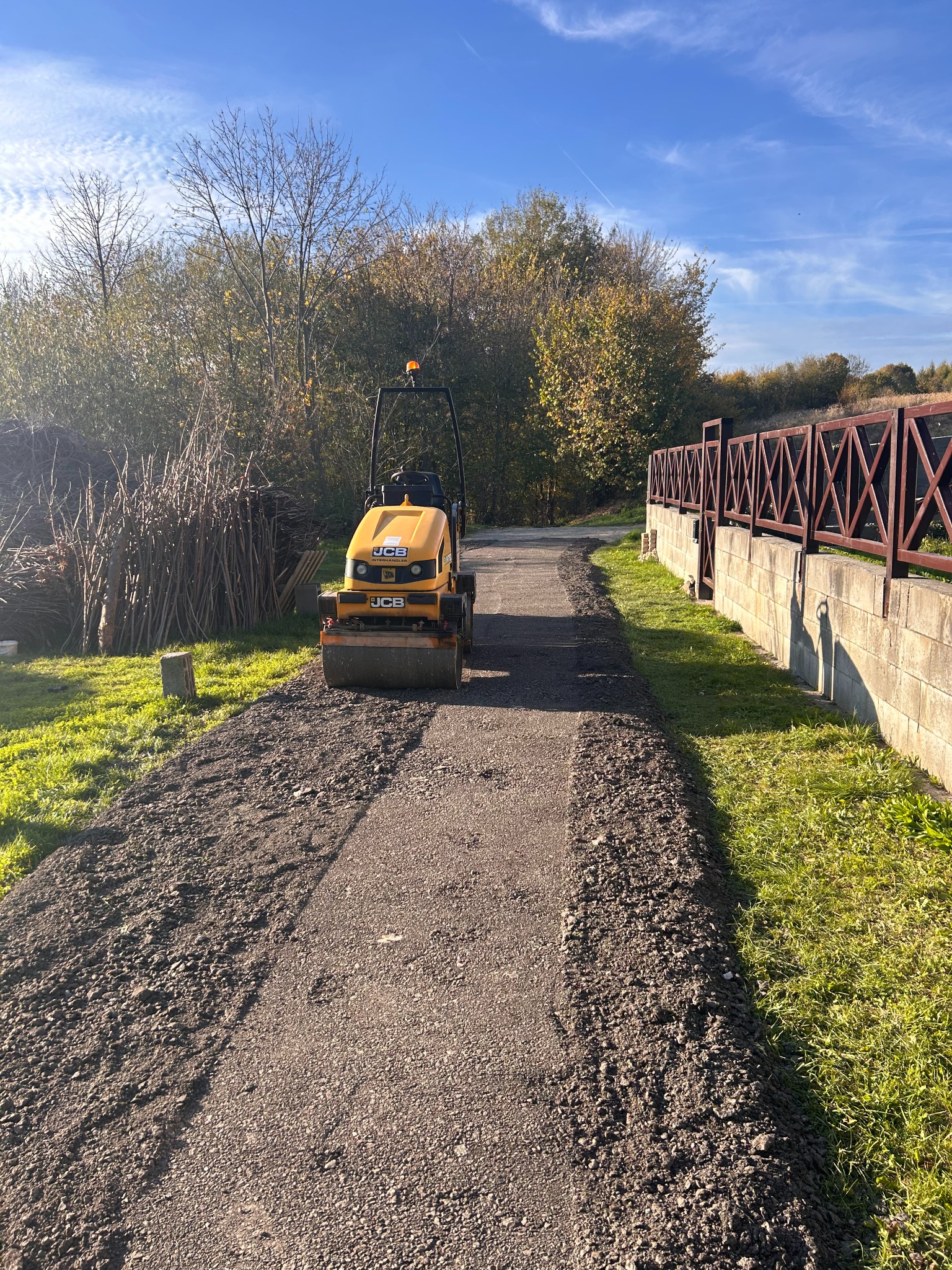 Żółty walec drogowy JCB ubija nawierzchnię ścieżki na tle zieleni i błękitnego nieba. Widoczne świeżo utwardzone fragmenty drogi.