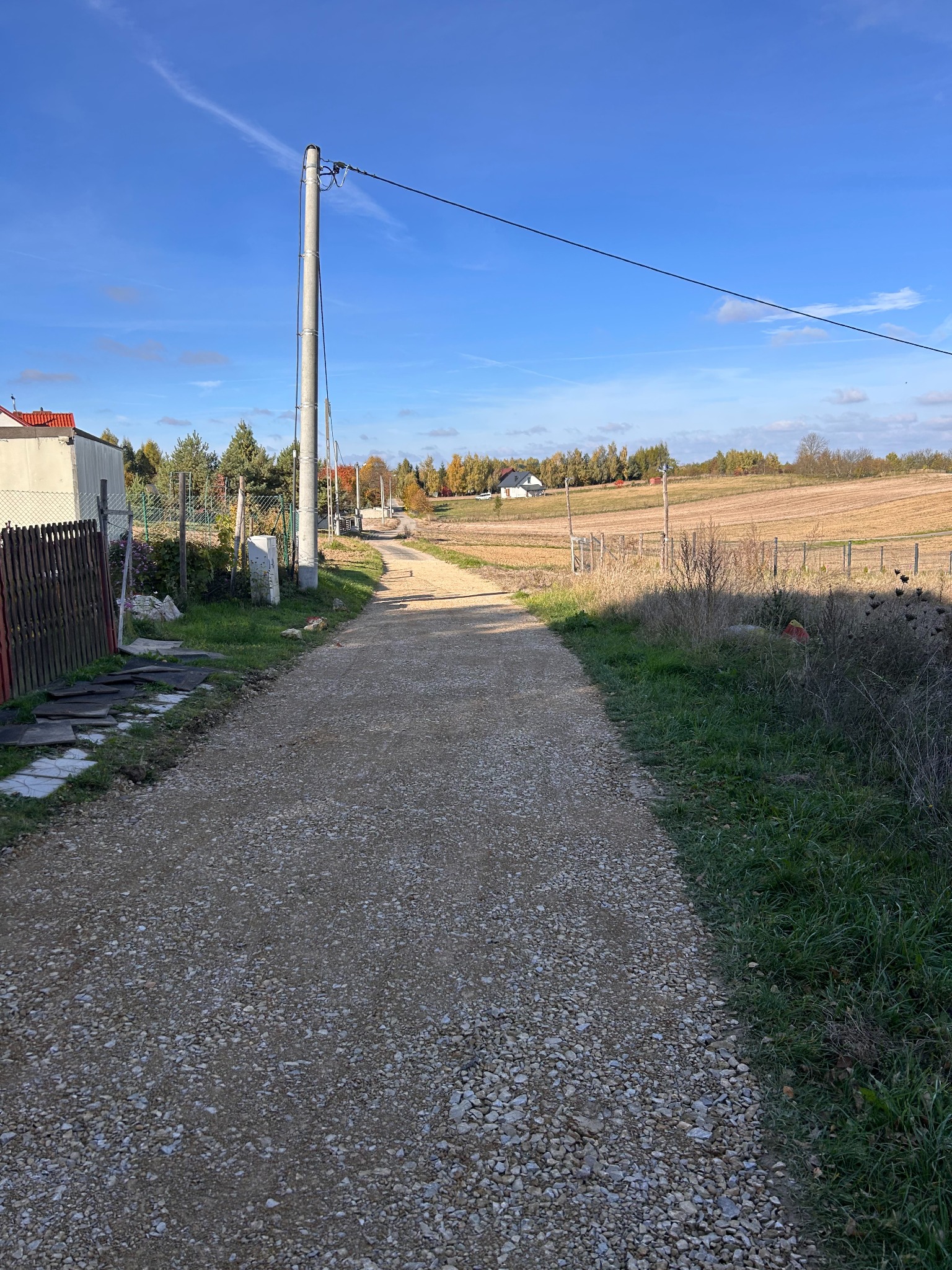 Utwardzona żwirowa droga wiejska w słoneczny dzień, widok z perspektywy pieszego, z budynkami i polami w tle.