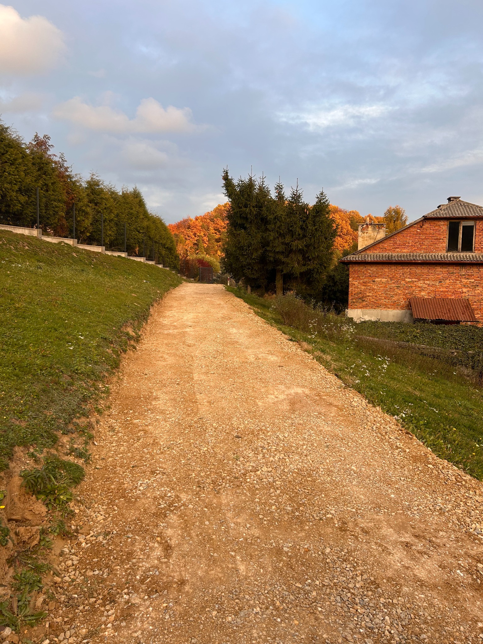 Żwirowa droga po utwardzeniu, widok z dołu. Po prawej stronie budynek z czerwonej cegły, w tle drzewa w jesiennych kolorach. Pochmurne niebo.