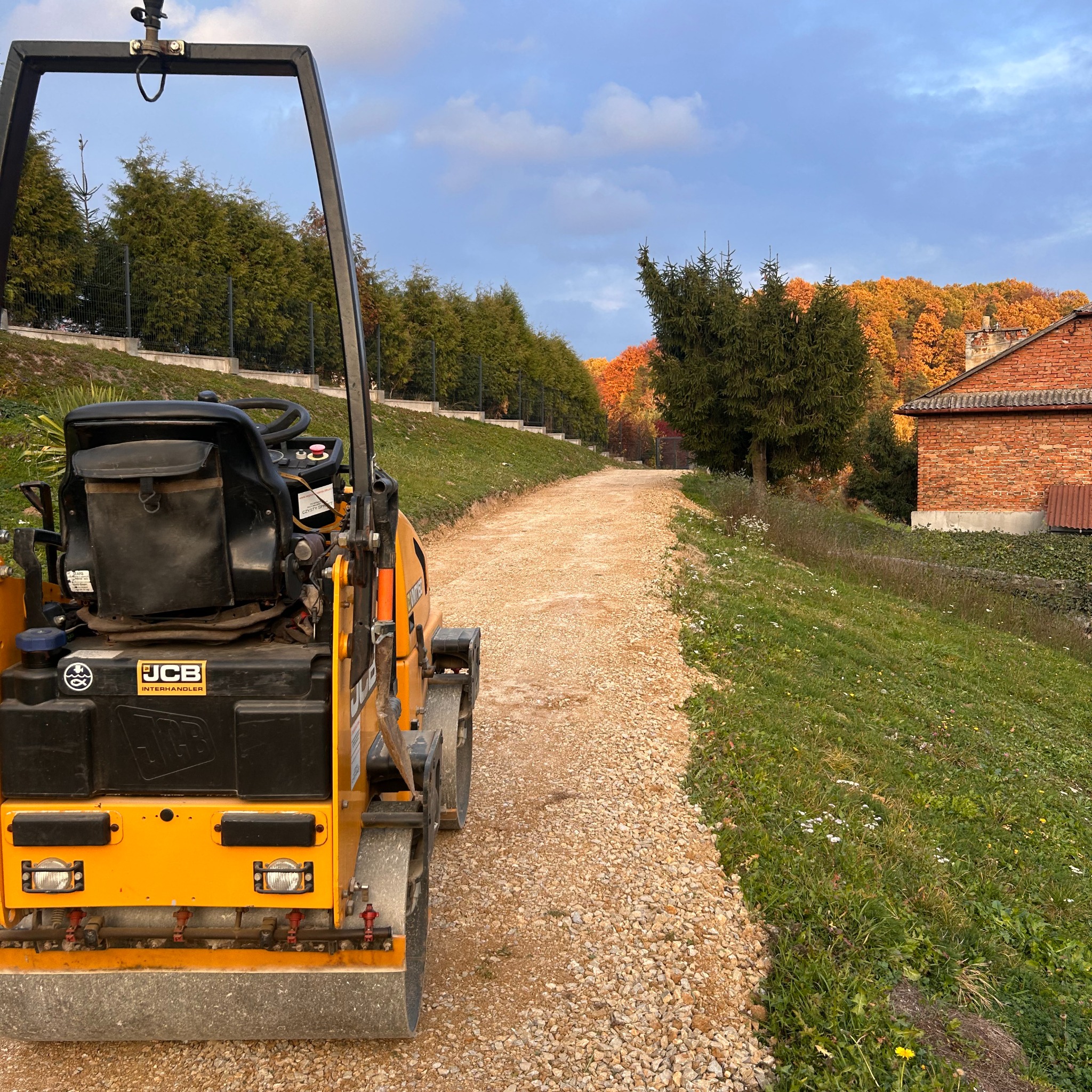 Żółty walec drogowy JCB ubija kamienistą ścieżkę na tle zieleni i ceglanego budynku. Widok z tyłu maszyny, jesienna sceneria.