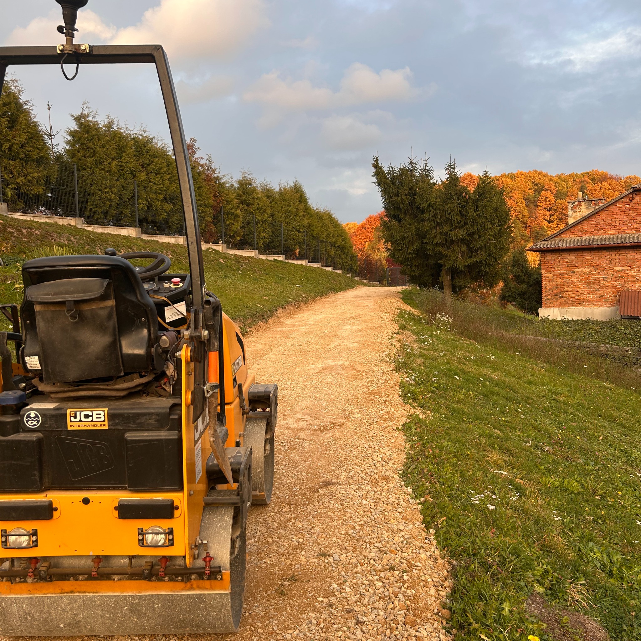 Żółty walec drogowy JCB ubija żwirową drogę gruntową na tle zieleni i jesiennych drzew, z budynkiem w tle. Prace ziemne, utwardzanie nawierzchni.