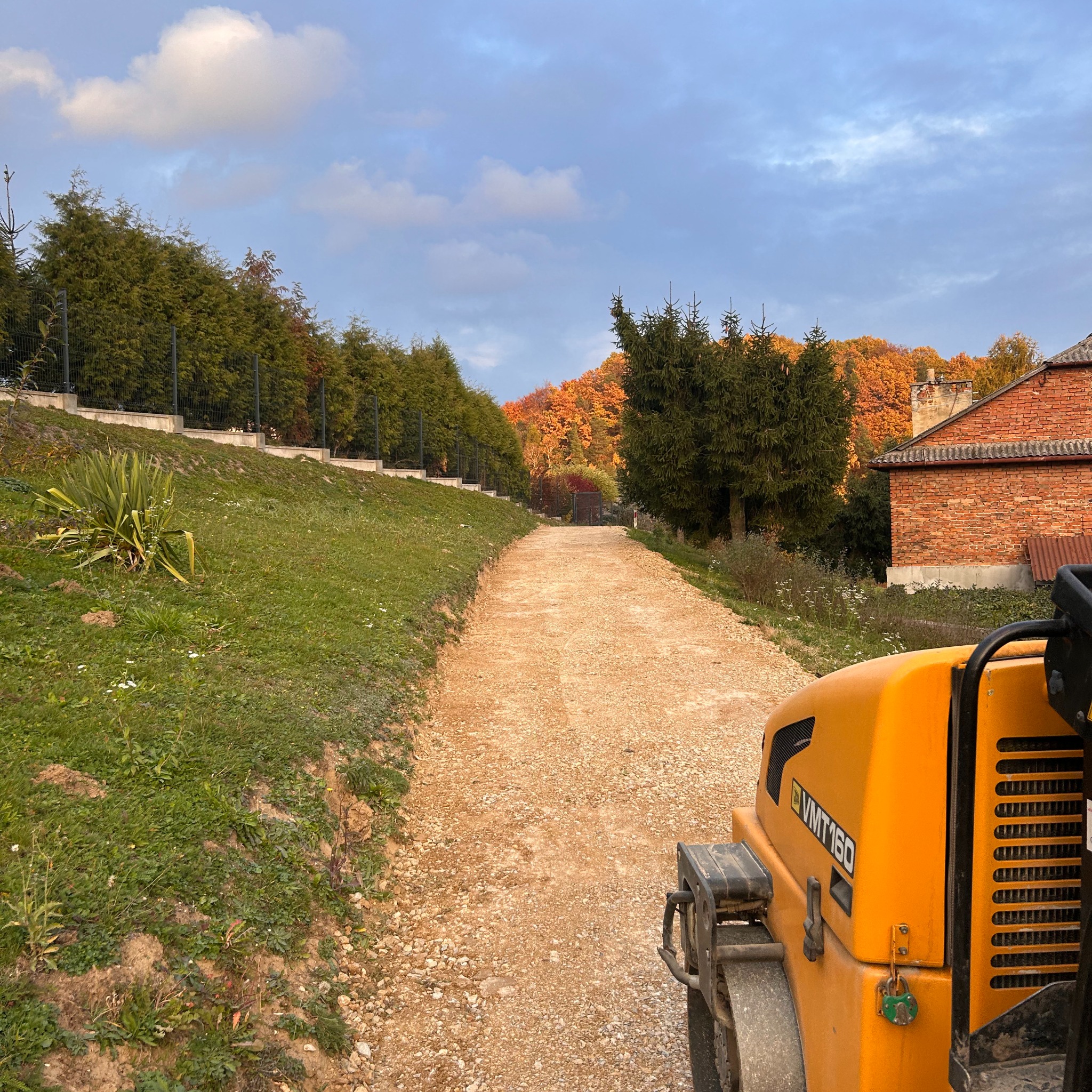 Żółty walec VMT160 na żwirowej drodze, w tle zieleń i budynek z czerwonej cegły. Widok z perspektywy operatora, w słoneczny dzień.