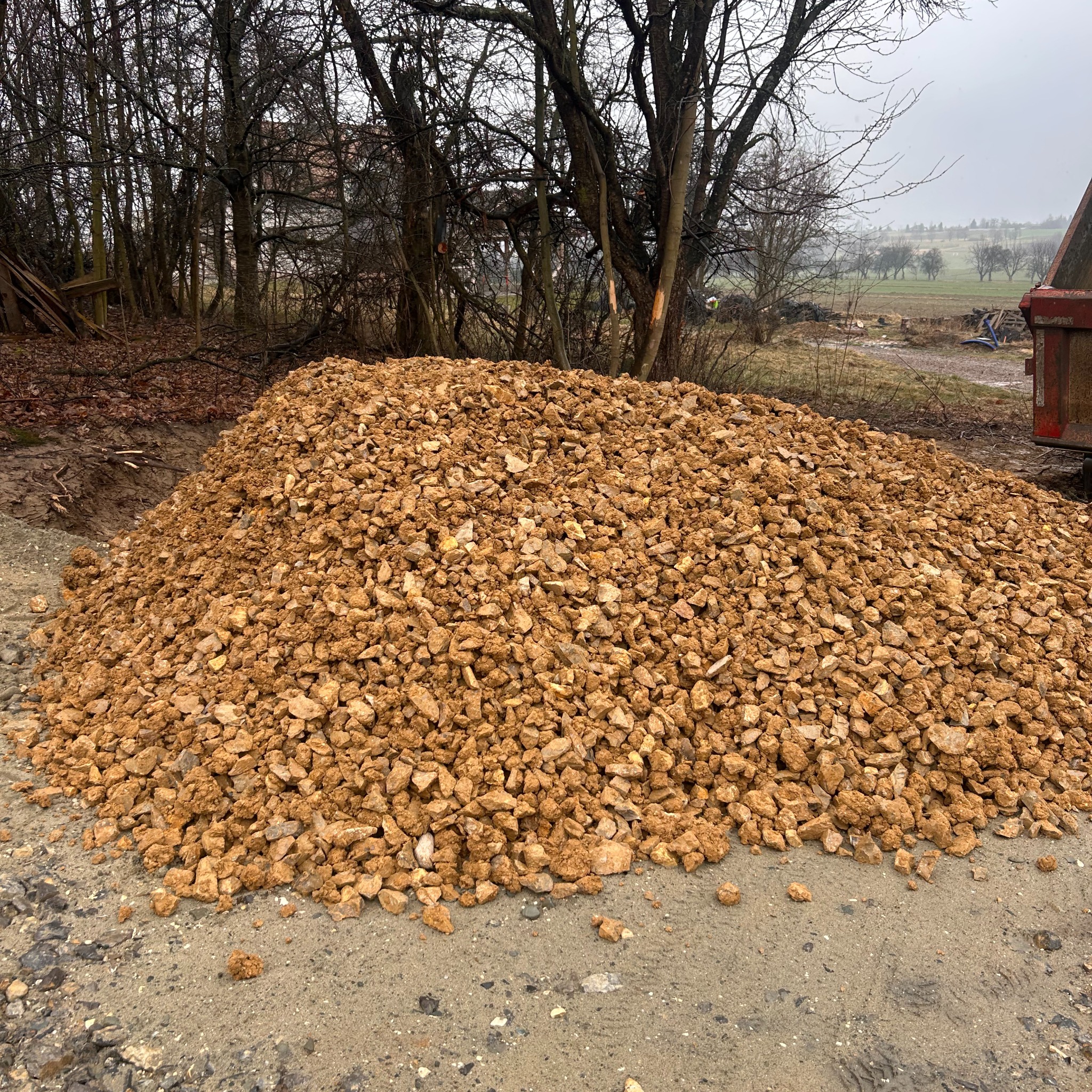 Kupa gruzu na utwardzonej drodze, w tle drzewa i pole. Fragment skrzyni ładunkowej pojazdu po prawej stronie kadru. Pochmurny dzień.