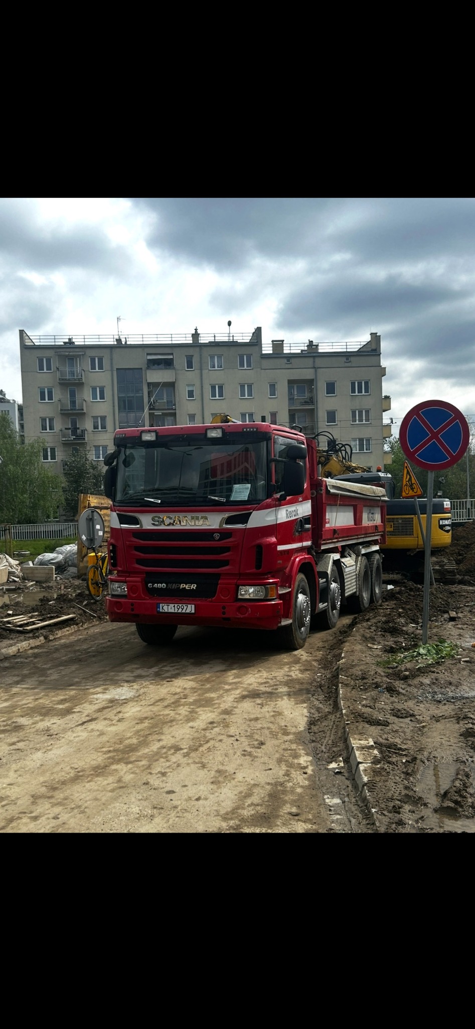 Czerwona ciężarówka Scania na placu budowy z koparką w tle, znak zakazu zatrzymywania się, budynek mieszkalny w tle. Prace ziemne w toku.