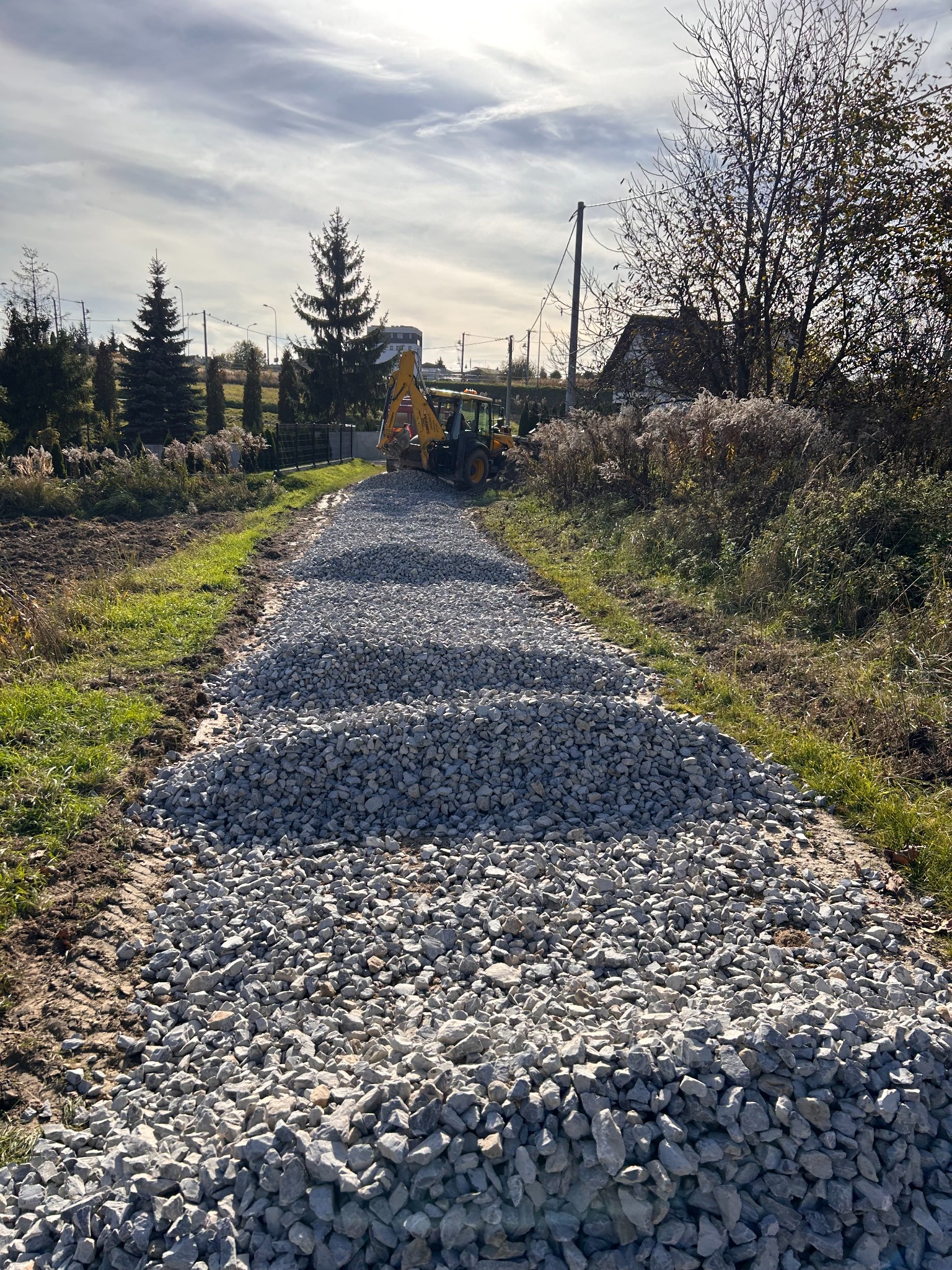 Żwirowa droga w trakcie utwardzania z widoczną koparką w oddali, otoczona zielenią i drzewami w słoneczny dzień. W tle domy i słupy energetyczne.