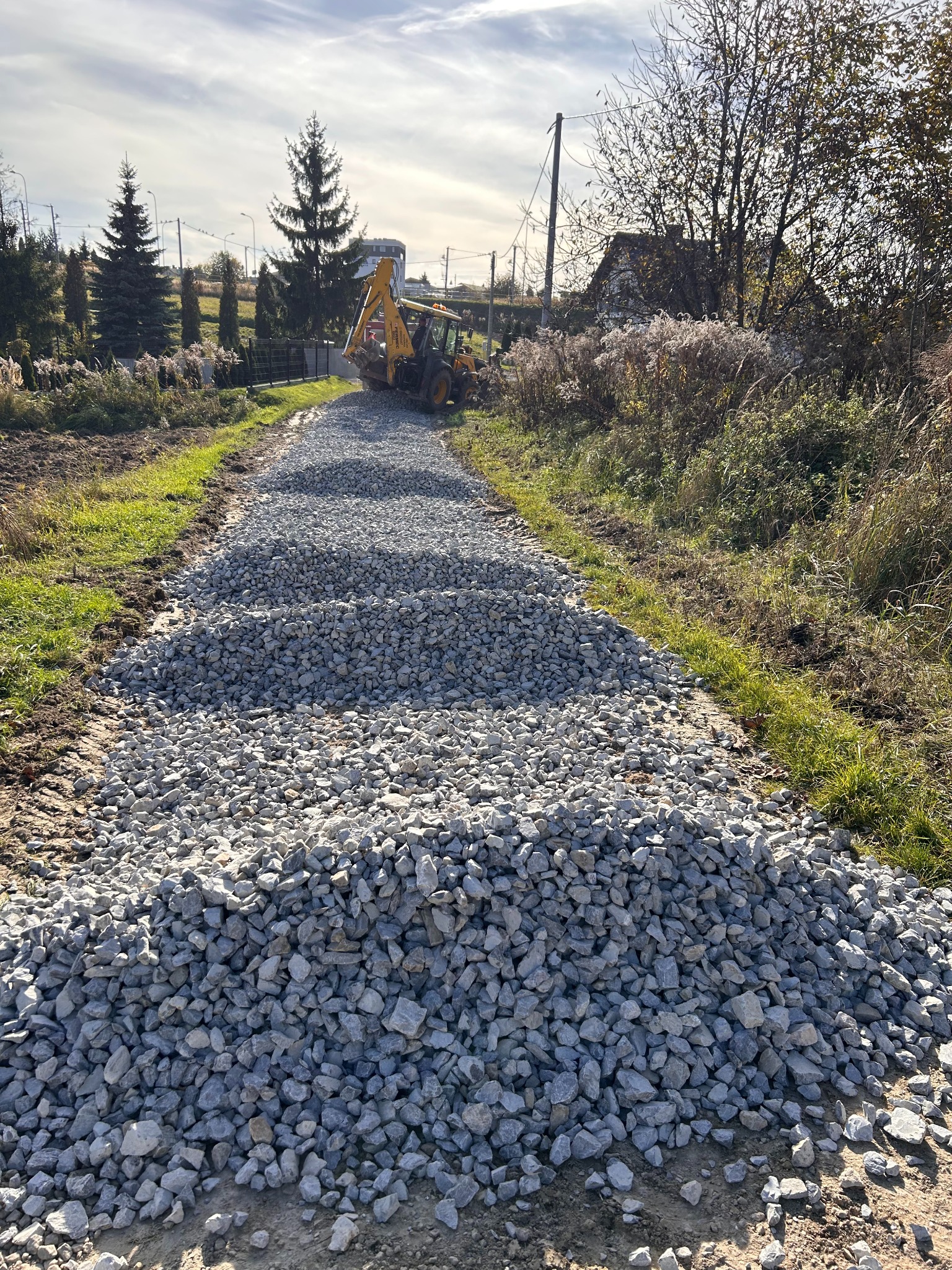 Żwirowa droga w trakcie budowy, z koparką w tle. Widoczne rozłożone kamienie i krajobraz wiejski w słoneczny dzień.