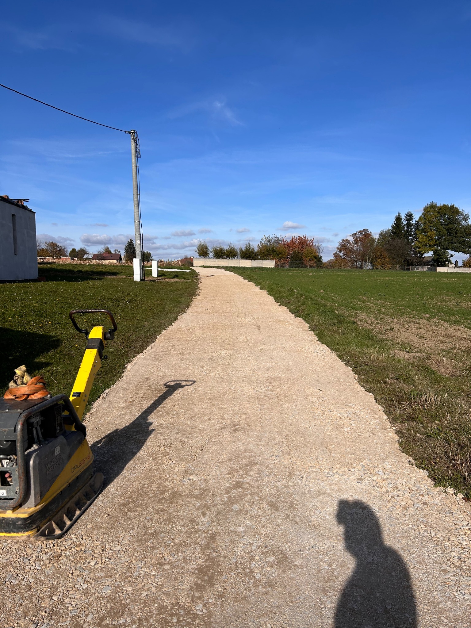 Utwardzona, żwirowa droga przez pole, z ubijarką Wacker Neuson na pierwszym planie i cieniem osoby; błękitne niebo w tle. Słoneczny dzień.