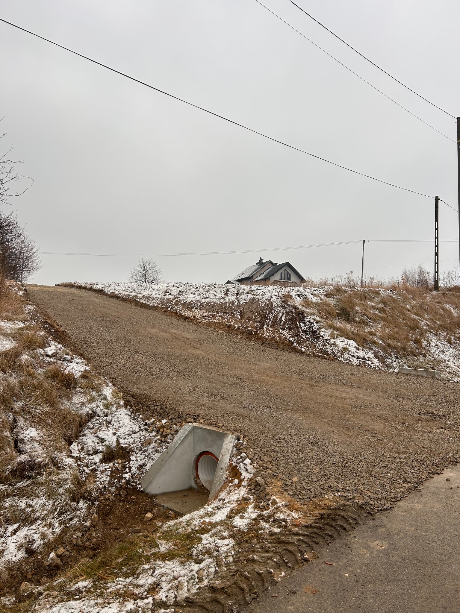 Utwardzona droga gruntowa na wzniesieniu z przepustem betonowym. Ślady opon, częściowo pokryta śniegiem. W tle dom i słupy energetyczne pod pochmurnym niebem.