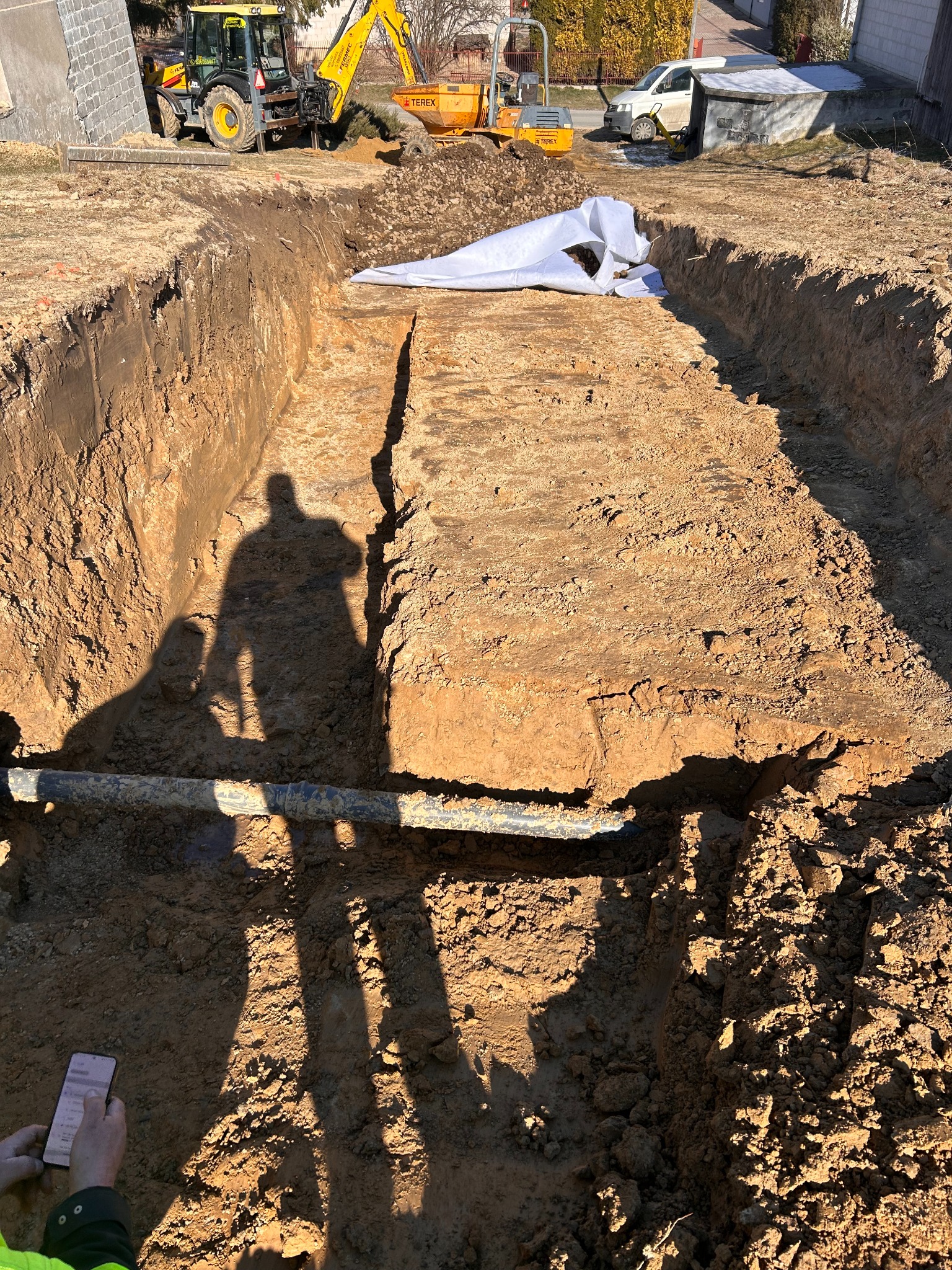 Długi wykop w ziemi z rurą na dnie, maszyny budowlane w tle. Cień osoby z telefonem na pierwszym planie. Wykop zabezpieczony folią.