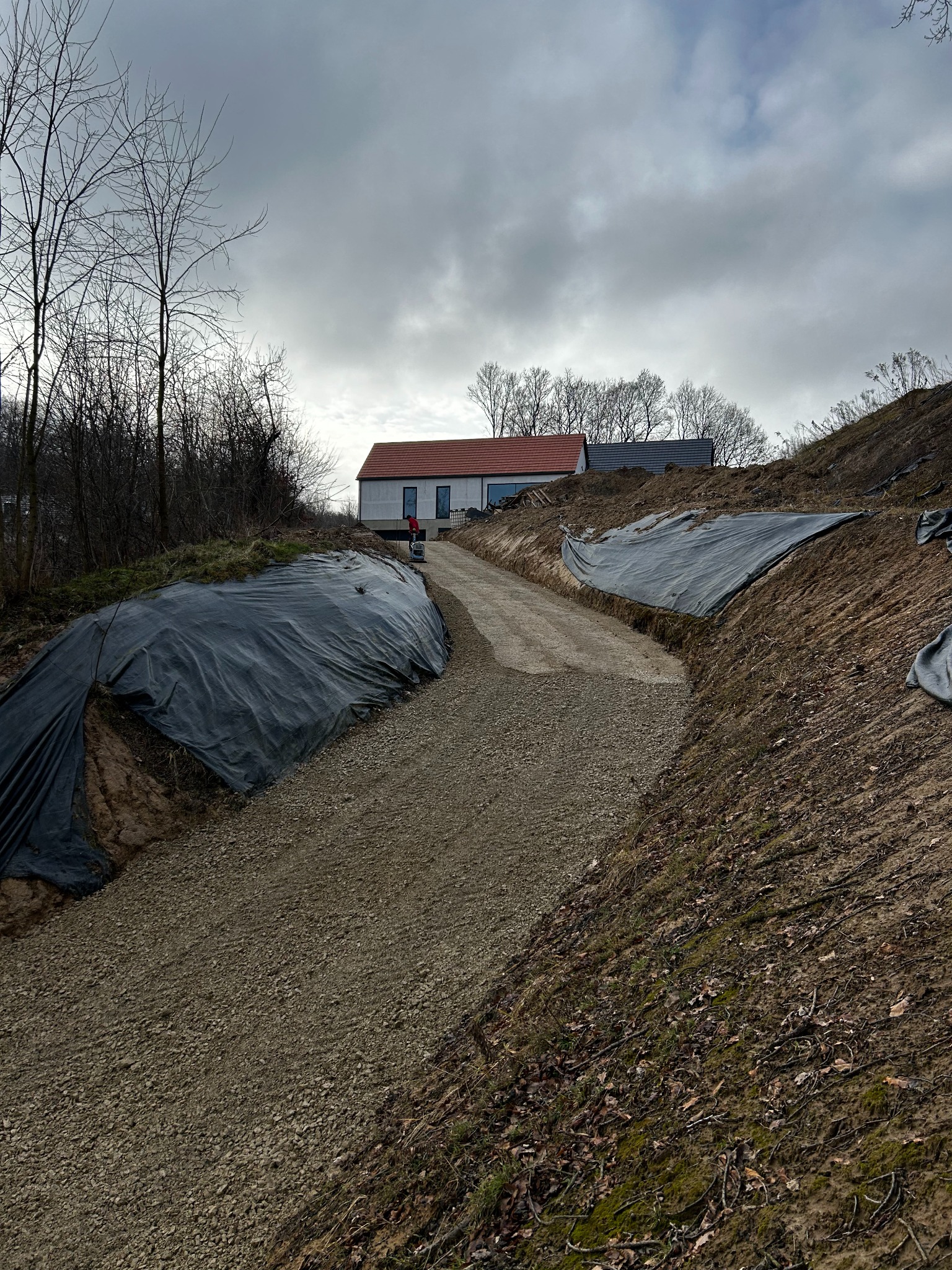 Utwardzona droga dojazdowa na wzgórzu, zabezpieczona czarną folią. W tle dom z czerwoną dachówką i pracownik z maszyną. Pochmurne niebo.