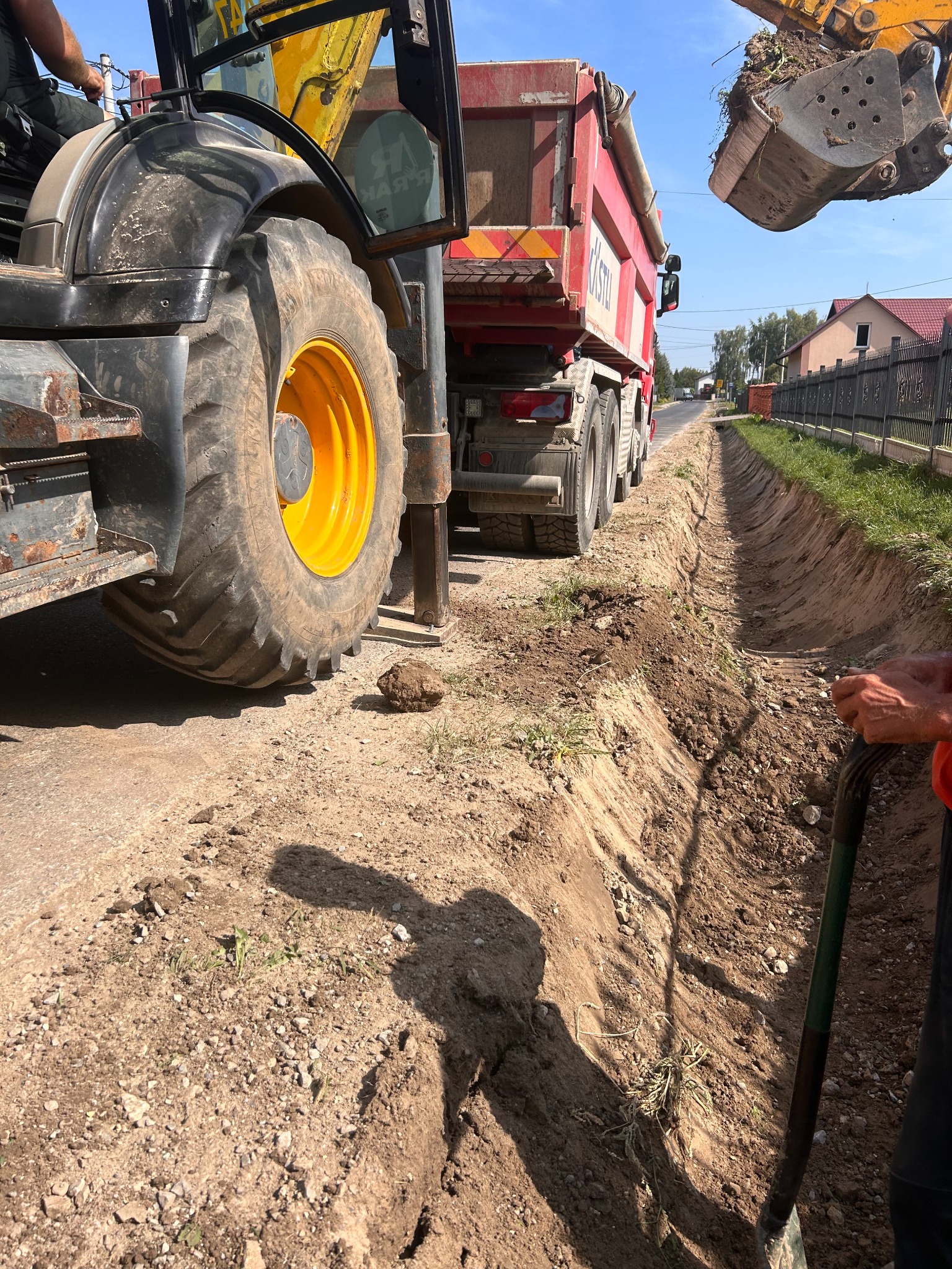 Koparka i wywrotka przy wykopie rowu wzdłuż drogi. Widoczny fragment koparki, koło traktora, wywrotka i rów. Prace ziemne w trakcie realizacji.