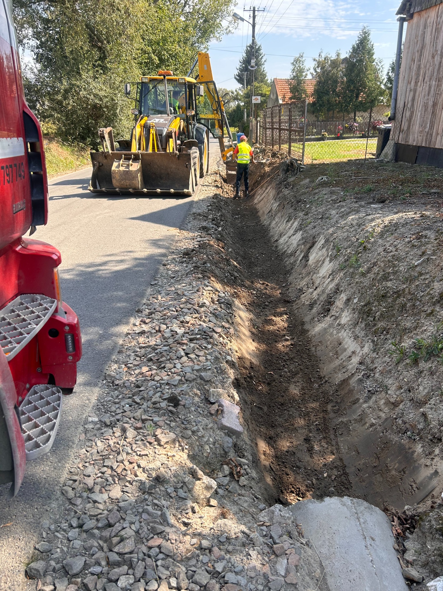 Żółta koparko-ładowarka JCB i robotnik w kamizelce przy wykopie w ziemi. W tle zabudowania i drzewa. Widoczny fragment czerwonej ciężarówki.