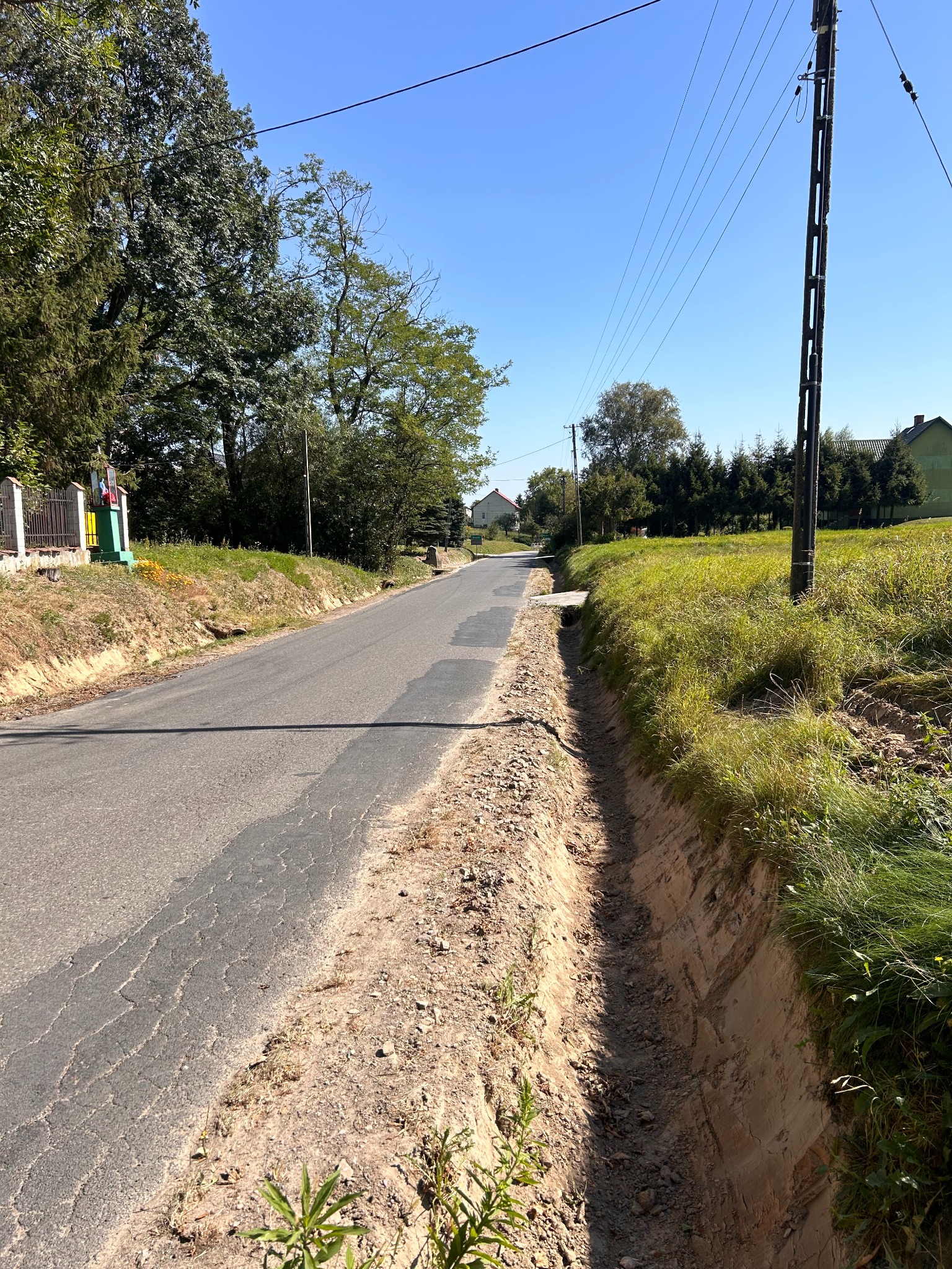 Długa, asfaltowa droga wiejska z rowem melioracyjnym po prawej stronie. Widoczne słupy elektryczne i zieleń. Niebo bezchmurne.