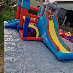 Fabryka Uśmiechu i Mebli David Parma - Kolorowy, dmuchany zamek ze zjeżdżalnią na szarej plandece. Dziecko zjeżdża. W tle budynki i zieleń. Widoczne detale konstrukcji i żywe kolory atrakcji.