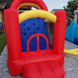 Kolorowy, dmuchany zamek z siatką ochronną na trawie, z widocznym fragmentem niebieskiej zjeżdżalni w tle. Zamek w odcieniach czerwieni, żółci i niebieskiego.