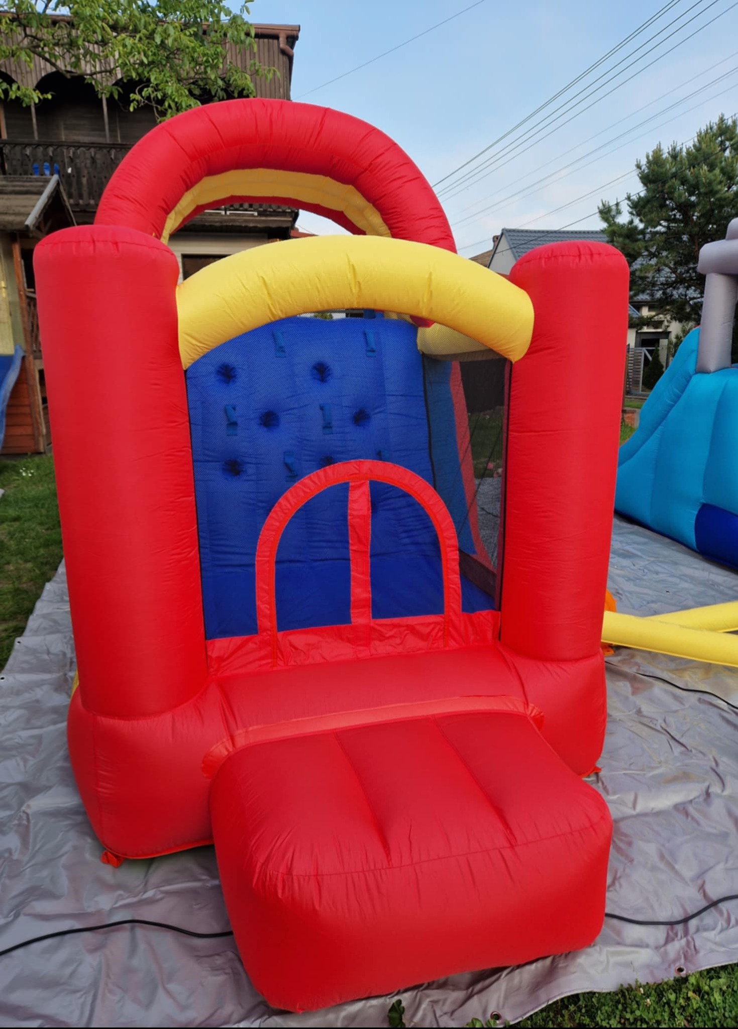 Kolorowy, dmuchany zamek z siatką ochronną na trawie, z widocznym fragmentem niebieskiej zjeżdżalni w tle. Zamek w odcieniach czerwieni, żółci i niebieskiego.