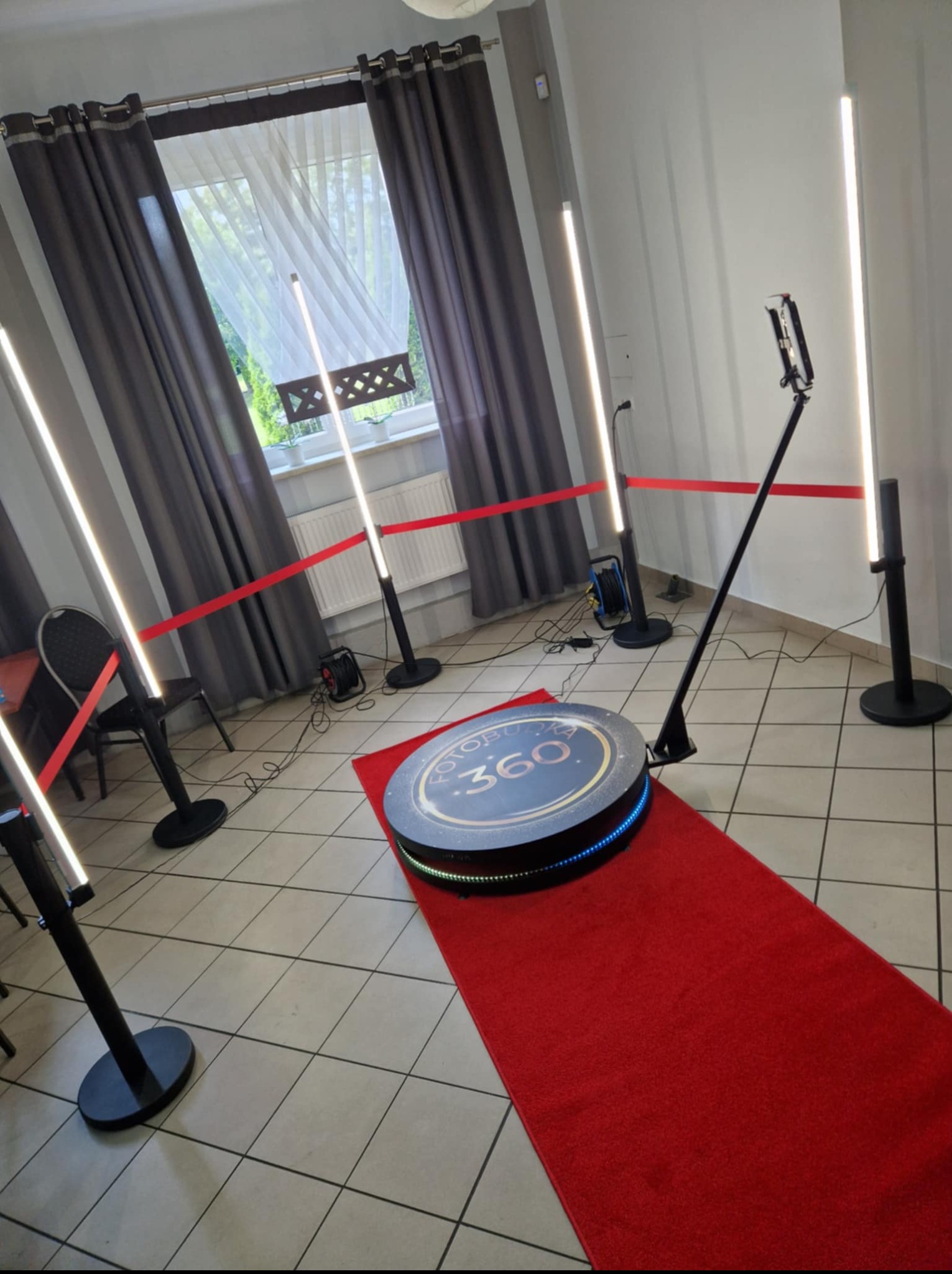 Rozstawiona fotobudka 360 z oświetleniem LED i czerwonym dywanem, przygotowana do użycia. W tle okno z szarymi zasłonami i białą firanką.