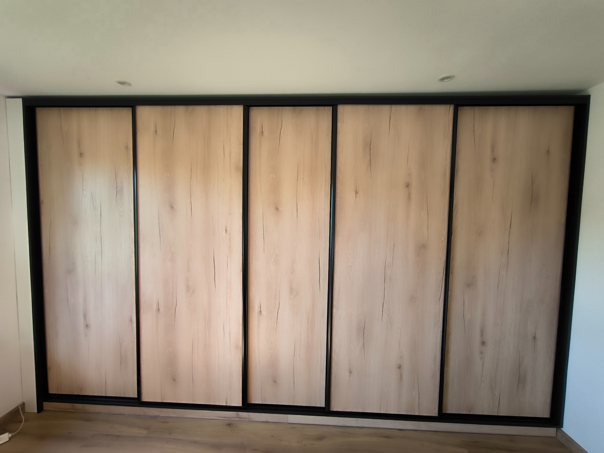 Duża szafa wnękowa z przesuwnymi drzwiami o jasnym, drewnopodobnym wykończeniu i czarnych obramowaniach, oświetlona punktowo, na tle jasnych ścian.