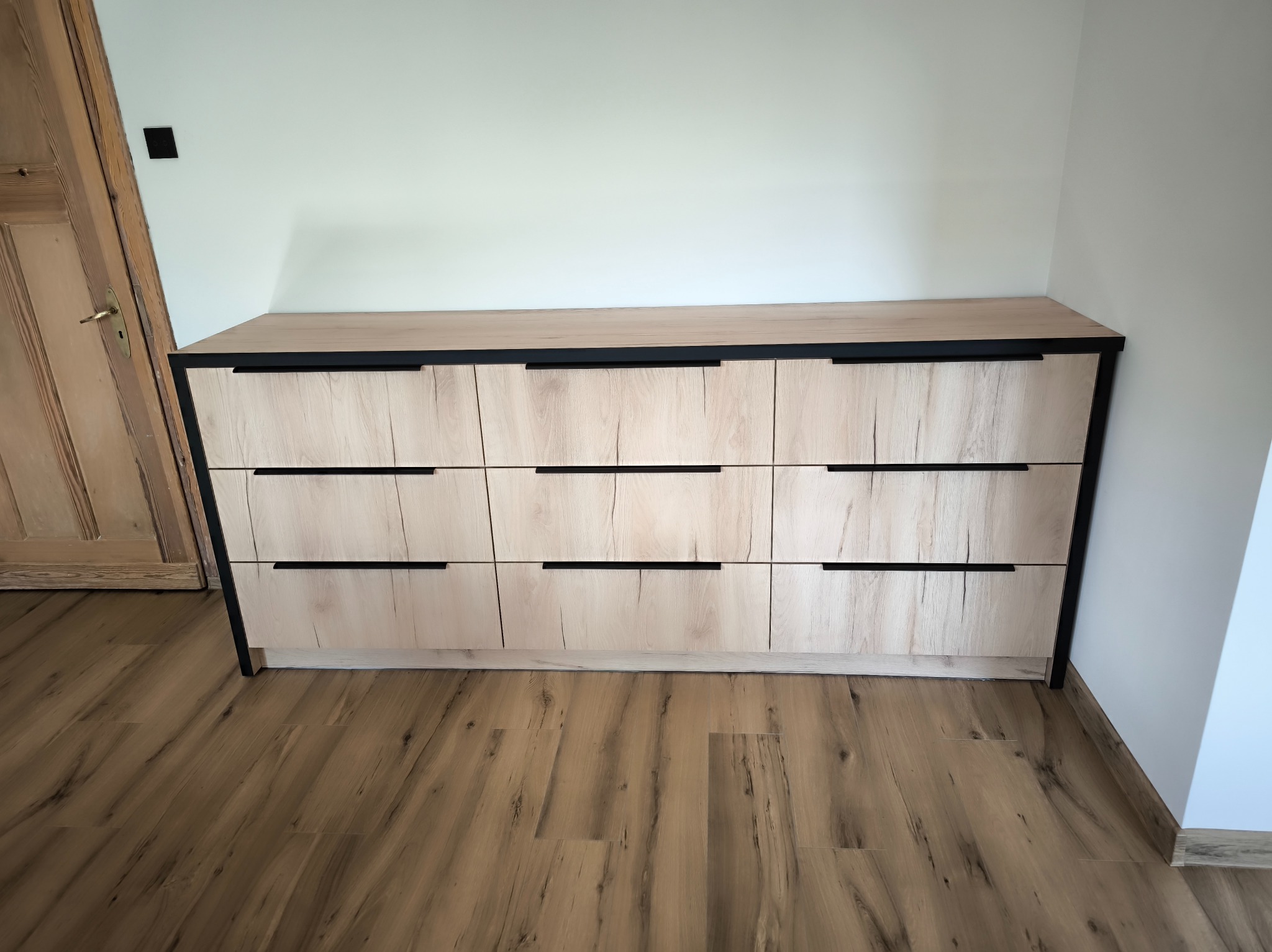 Komoda z jasnego drewna z czarnymi uchwytami, trzy rzędy szuflad, nowoczesny design, widok z przodu na tle białej ściany i drewnianej podłogi.