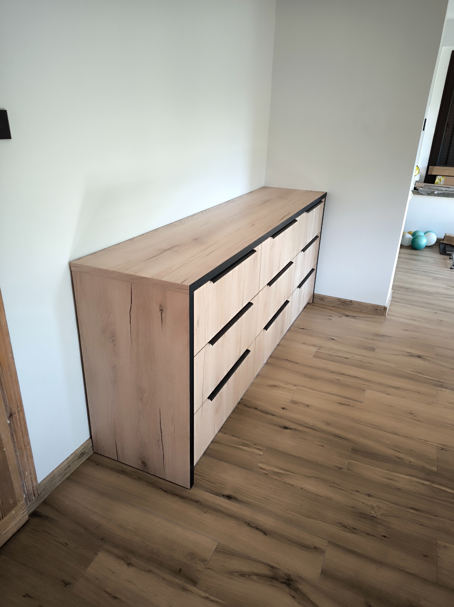 Jasna komoda z czarnymi uchwytami, drewnopodobny dekor, ustawiona w rogu pokoju z panelami podłogowymi. Minimalistyczny design.