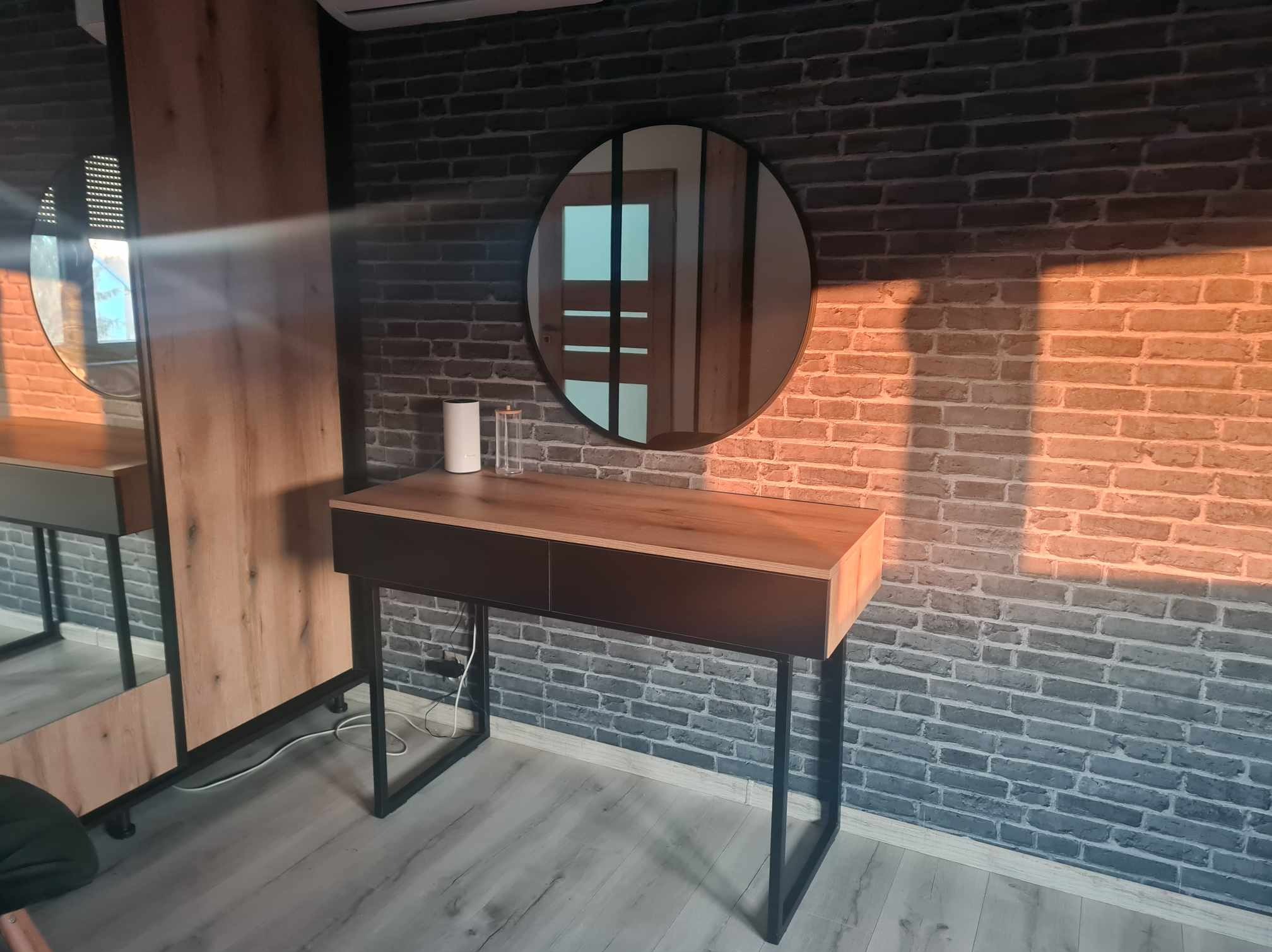 Nowoczesna toaletka z okrągłym lustrem na tle ceglanej ściany. Blat w odcieniach drewna, czarne metalowe nogi. Minimalistyczny design, ciepłe oświetlenie.