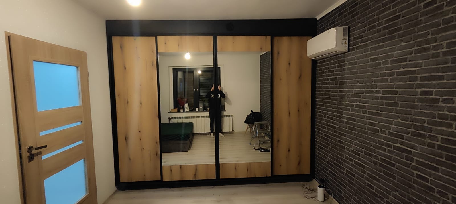 Nowoczesna szafa przesuwna z lustrami i drewnianymi panelami, czarne obramowanie, ściana z cegły, klimatyzator. Widoczny fragment drzwi z przeszkleniem.