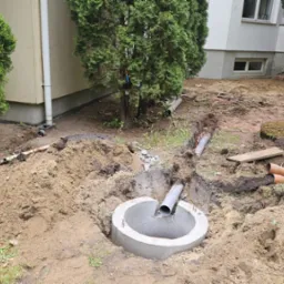 Montaż rur kanalizacyjnych w wykopie na zewnątrz budynku. Widoczny betonowy krąg i rury z tworzywa sztucznego, ziemia oraz narzędzia pracy.