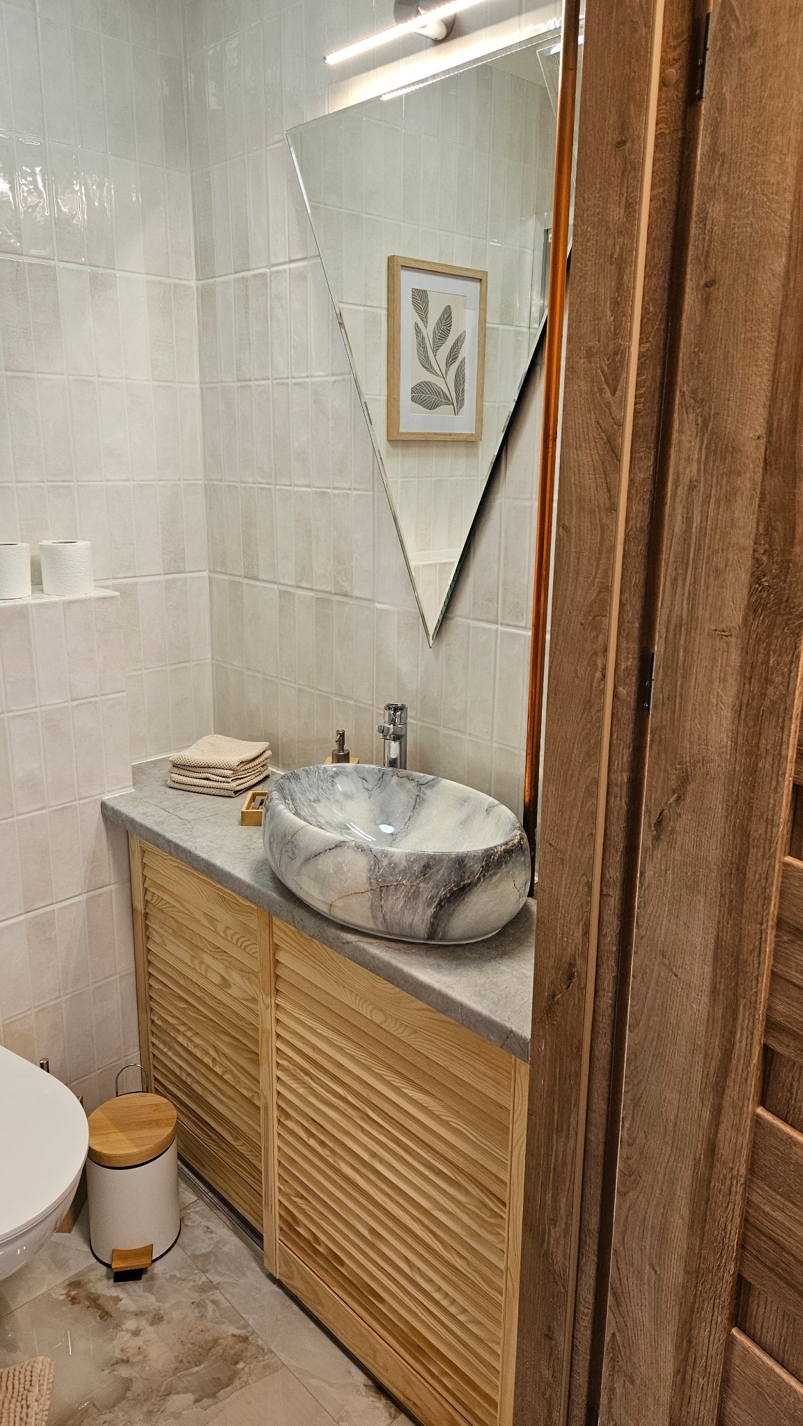 Stylowa łazienka z marmurową umywalką na drewnianej szafce, lustro w kształcie trapezu, białe płytki na ścianach i drewniane drzwi. Ujęcie z bliska.