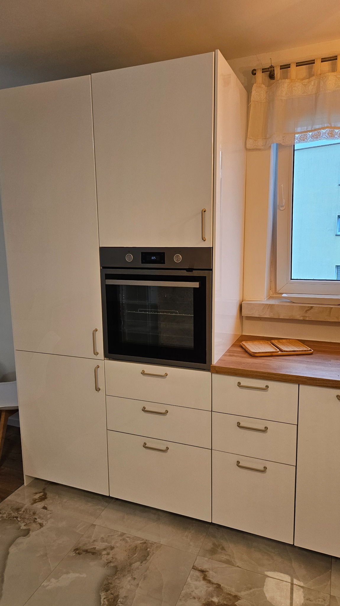 Nowoczesna zabudowa kuchenna z piekarnikiem w kolorze białym, z drewnianym blatem i jasnymi uchwytami. Widok na okno z firanką.