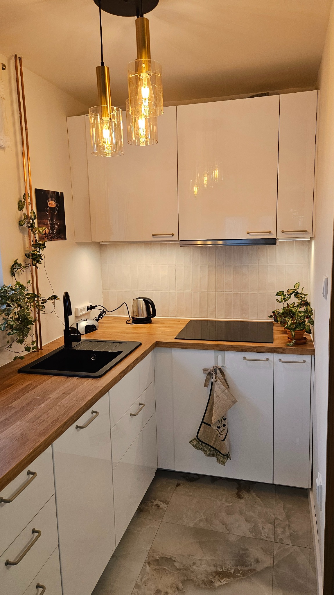 Nowoczesna kuchnia w bieli z drewnianym blatem i złotymi akcentami oświetleniowymi. Rośliny dodają świeżości, a marmurowa podłoga elegancji.