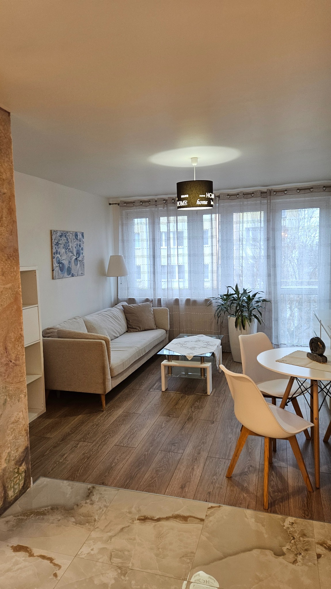 Jasny salon z drewnianą podłogą, sofą, stolikiem kawowym i okrągłym stołem z krzesłami. Lampa wisząca z napisem 'Home' dodaje przytulnego charakteru.