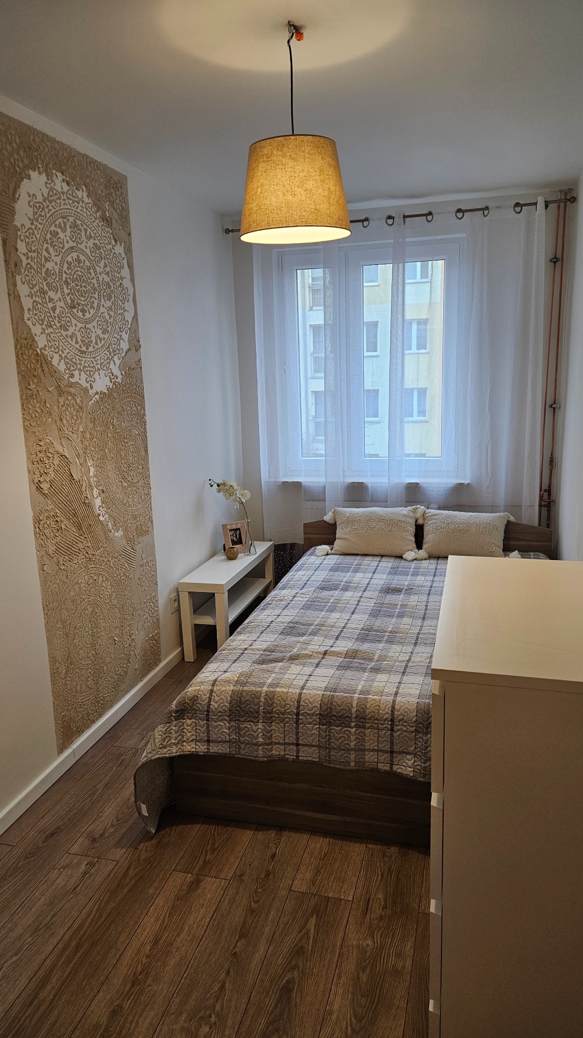 Przytulna sypialnia po remoncie: dekoracyjna ściana z ornamentem, drewniana podłoga, łóżko z wzorzystą narzutą i lampa wisząca. Nowoczesny design.