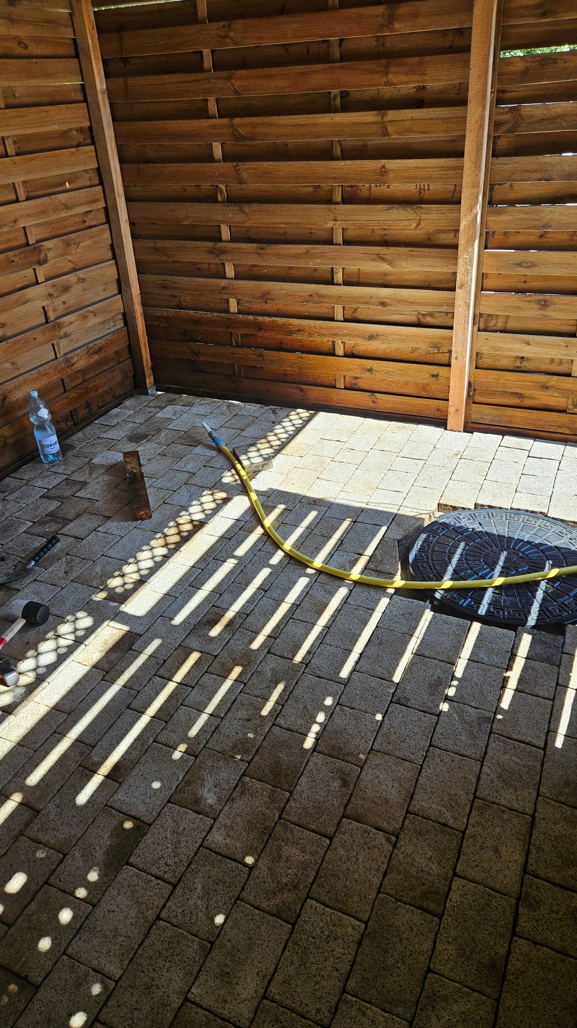Układanie kostki brukowej w drewnianej altanie. Wąż ogrodowy, narzędzia i butelka wody obok. Promienie słońca tworzą wzory na kostce.