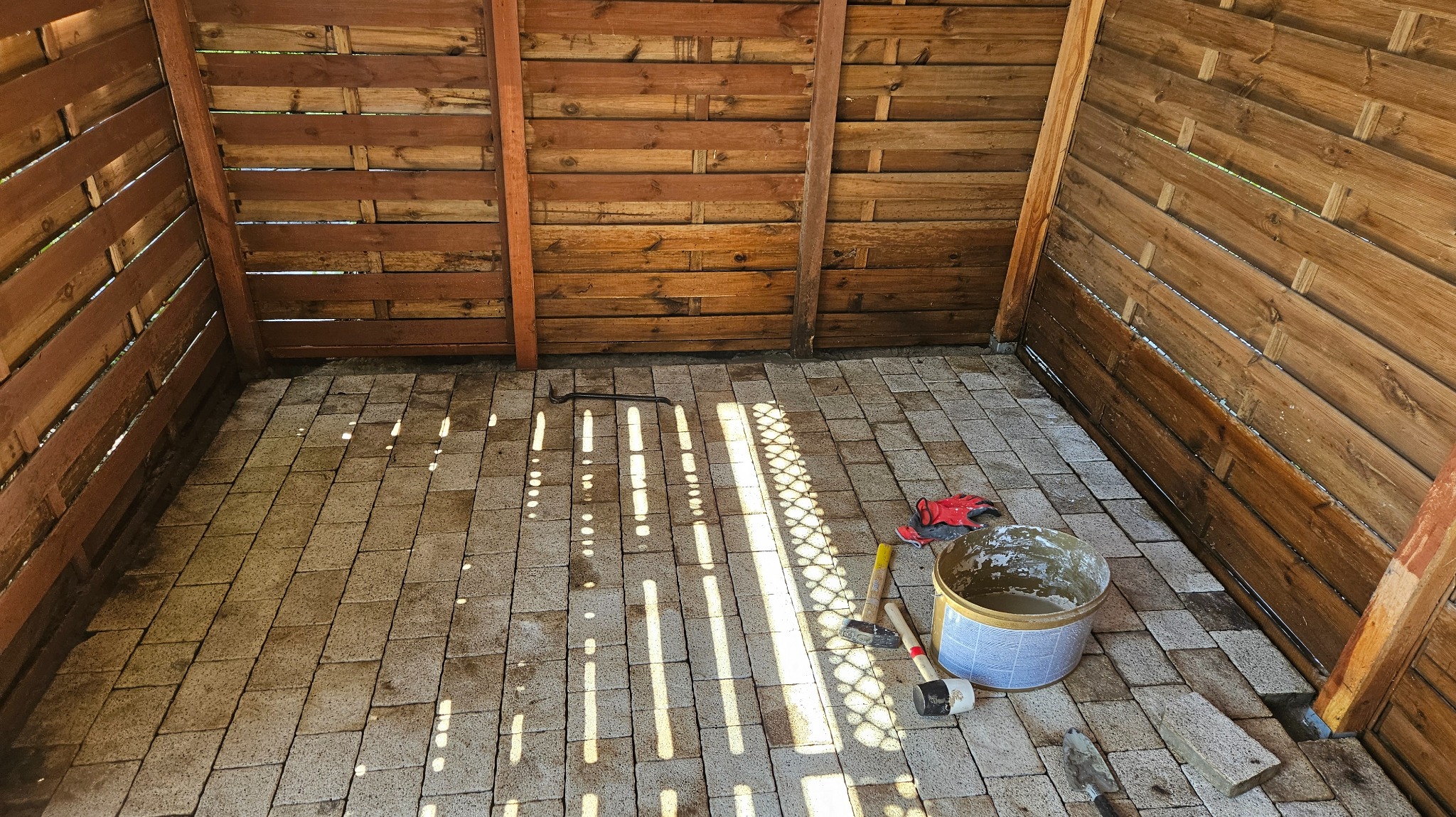 Drewniana altana z nową, ułożoną kostką brukową na podłodze. Widoczne narzędzia: młotek, łom, wiadro z zaprawą i rękawice robocze. Promienie słoneczne wpadają do środka.